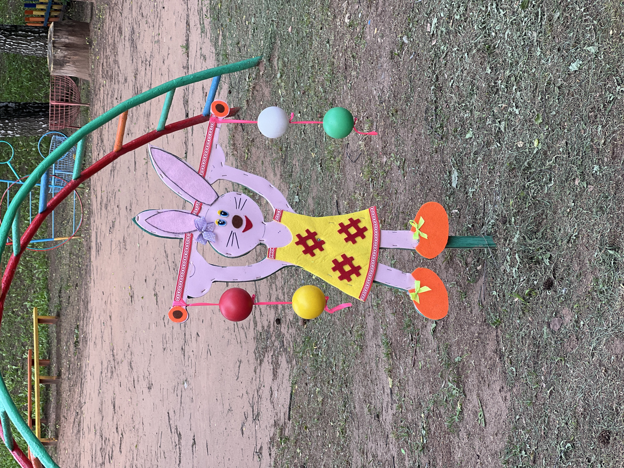 In the city of Советско-Никольское, Russia, a pink cutout of a cartoon bunny stands as a decorative element on a playground. The bunny, adorned in a yellow dress with red hash symbols, is positioned outdoors on a dirt and grass ground. It holds a pink ribbon in its outstretched arms, from which red, yellow, white, and green balls are suspended. The bunny's feet are represented by orange ovals with green bows. In the background, a colorful jungle gym structure with green, red, and blue bars dominates the scene. Beyond the playground equipment, a few trees with white trunks are visible, along with a wooden stump and a short fence. The ground is a mix of sandy soil and sparse, dry grass, with some scattered pinecones.