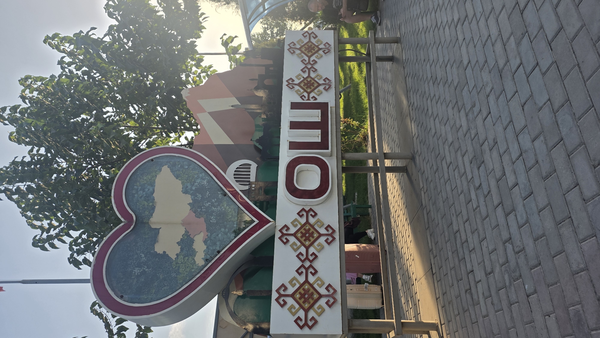 In the foreground, a brick-paved pathway is visible. Behind the pathway, a bench with metal legs and a wooden seat is situated in front of a white decorative sign. The sign features Cyrillic letters, the word "ОШ," and intricate red and yellow patterns on either side. Above the sign, a large heart-shaped structure displays a map of Kyrgyzstan. The background includes lush green trees and a clear sky. On the right side of the image, a man is seated on a bench, looking at his phone. Two suitcases are placed near the left side of the decorative sign. A portion of a transparent, curved structure, possibly a bus stop shelter, is visible on the right. The image is taken outdoors in what appears to be a public space.