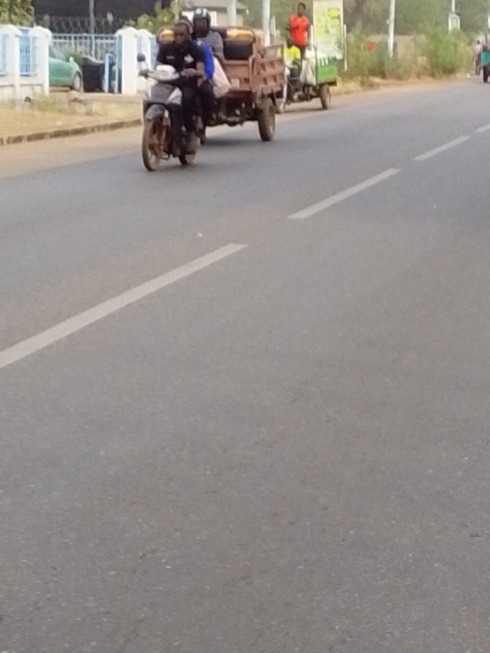 The scene is an asphalt road under daylight in the city of Unknown, Ghana. A dashed white line marks the road's center or lane division.
In the mid-ground, a dark-colored motorcycle pulls a two-wheeled wooden trailer, moving rightward. The motorcycle is occupied by two individuals wearing helmets. The driver wears a dark shirt, and the passenger wears a blue top. The wooden trailer carries brown cargo.
Further right and behind the motorcycle, a green and white motorized tricycle proceeds in the same direction. An individual in an orange shirt stands in the tricycle's rear section, with another person in a yellow shirt partially visible at the front.
The left roadside features a light-colored curb, a blue metal fence with a gate, a partial white building, and a green car parked behind the fence. The right roadside consists of an unpaved shoulder with sparse vegetation leading to distant trees.