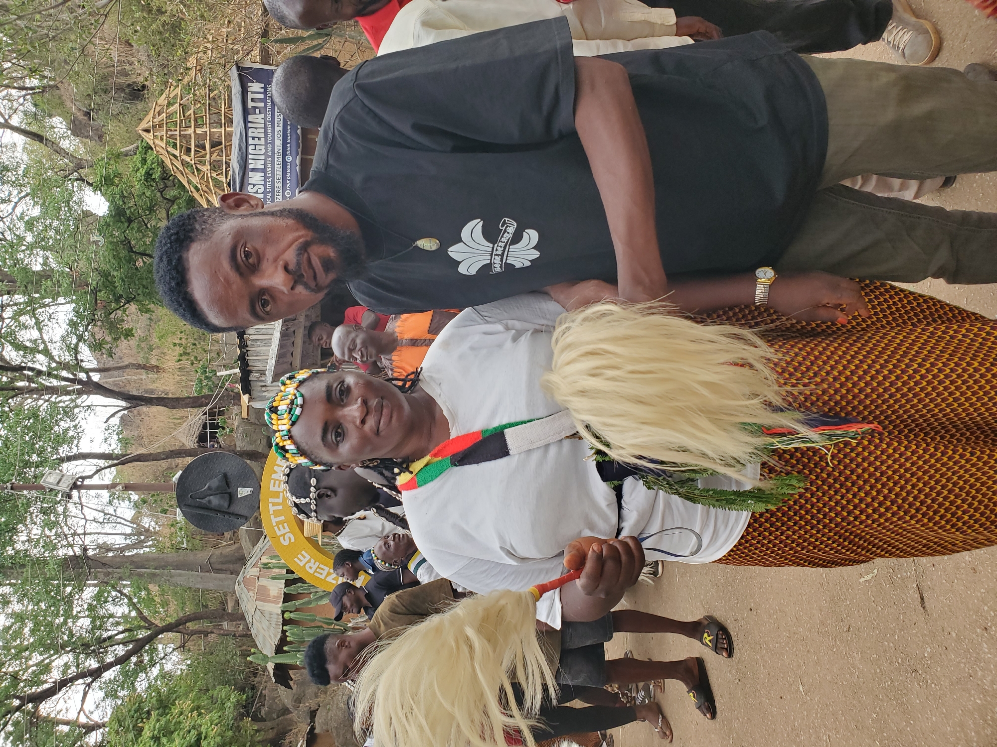 This outdoor, daylight image captures a cultural or tourism-related event, likely in Jos, Nigeria. In the foreground, a man and a woman are centered, looking towards the viewer. The man, with a beard, wears a black t-shirt featuring a white emblem with the text "HOME HEARTS" and a round pendant necklace. He is standing close to the woman, whose left wrist, adorned with a gold-colored watch, is visible.
The woman wears a white t-shirt with a colorful, striped fabric band (red, black, green, white) draped across her shoulder. She has a beaded headband in yellow, green, and black, and her hair is styled with braids. She holds a light-colored, whisk-like object, possibly made of hair or fibers, in her right hand. Her lower body is covered by a patterned skirt in shades of brown and orange. Another similar whisk-like object is partially visible in the bottom foreground, held by another person.
In the background, various people are gathered, and a natural setting with numerous trees is apparent. A wooden skeletal structure, possibly a traditional hut under construction or a decorative element, stands out among the trees. Prominent signage indicates the event's nature: "SM NIGERIA-TTN" (Tourism Nigeria), "TOURIST DESTINATIONS," and mentions "ZERE SETTLEMENT, JOS MUSEUM." A circular yellow sign also reads "ZERE SETTLEMENT." Further details on the sign include social media handles like "@tour_plateau" and "@think_tourism_n." Emotions appear generally relaxed, with the main subjects displaying neutral to slight smiles. The weather seems clear and bright.