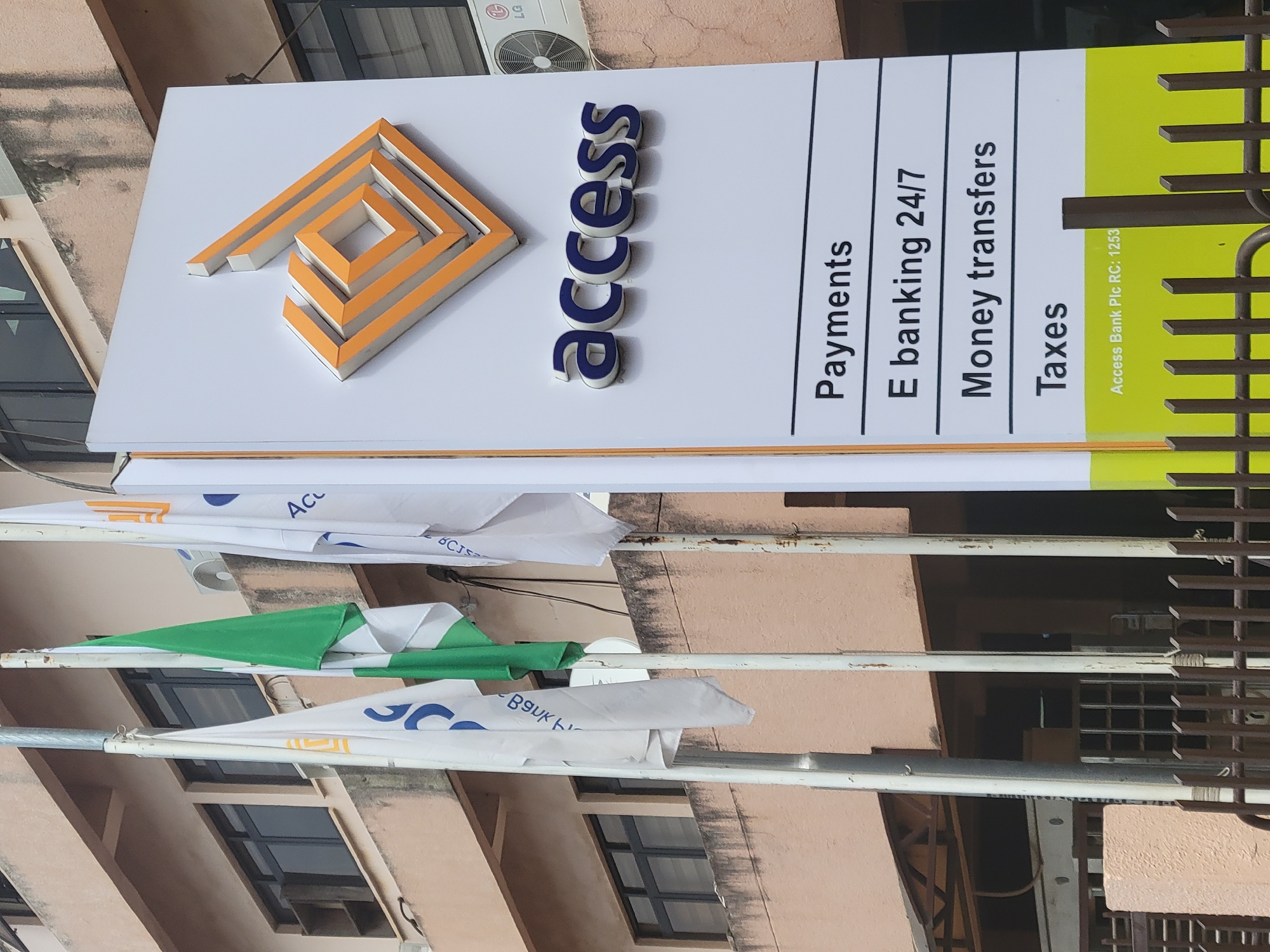 The image shows an Access Bank sign. The sign is white with the Access Bank logo and the word "access" in blue. Below that is the text "Payments", "E banking 24/7", "Money transfers", and "Taxes". The lower right corner of the sign is green and includes the text "Access Bank Plc RC: 1253". There are also flags on poles below the sign, with "ACL", "BgUKト", and "CIS" visible on the flags. The background shows a light-brown building with windows and an LG air conditioning unit. The presence of the Access Bank sign suggests this image was taken in a commercial area, potentially in Abuja, Nigeria.