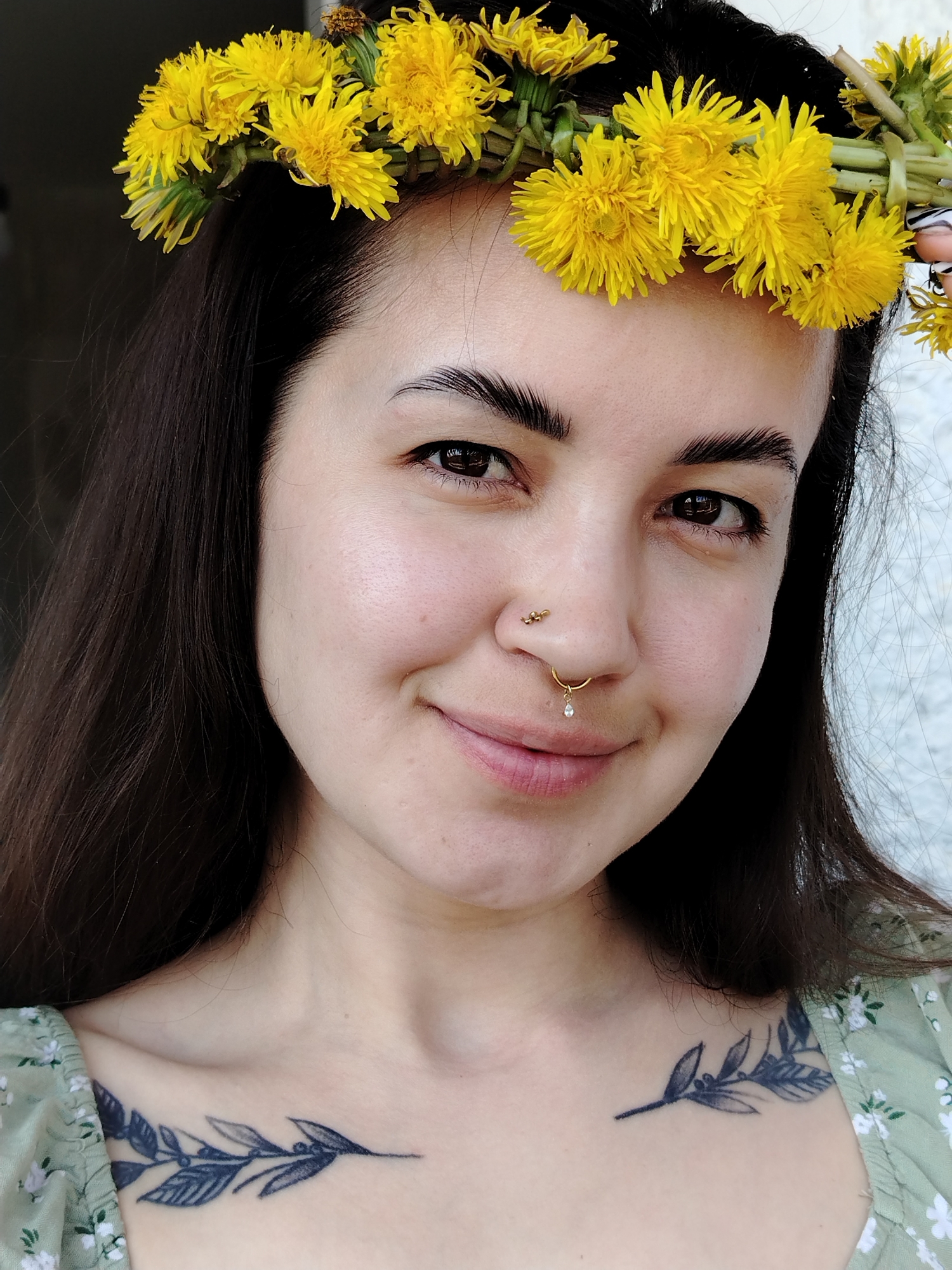 A young woman with dark hair is wearing a crown of yellow dandelions. She has a nose piercing on both sides of her nose. She is wearing a light green floral print top, and has a tattoo of a branch with leaves on her upper chest. The background appears to be a plain, light-colored wall. The image does not provide any information about the city of Noviy, Russia.