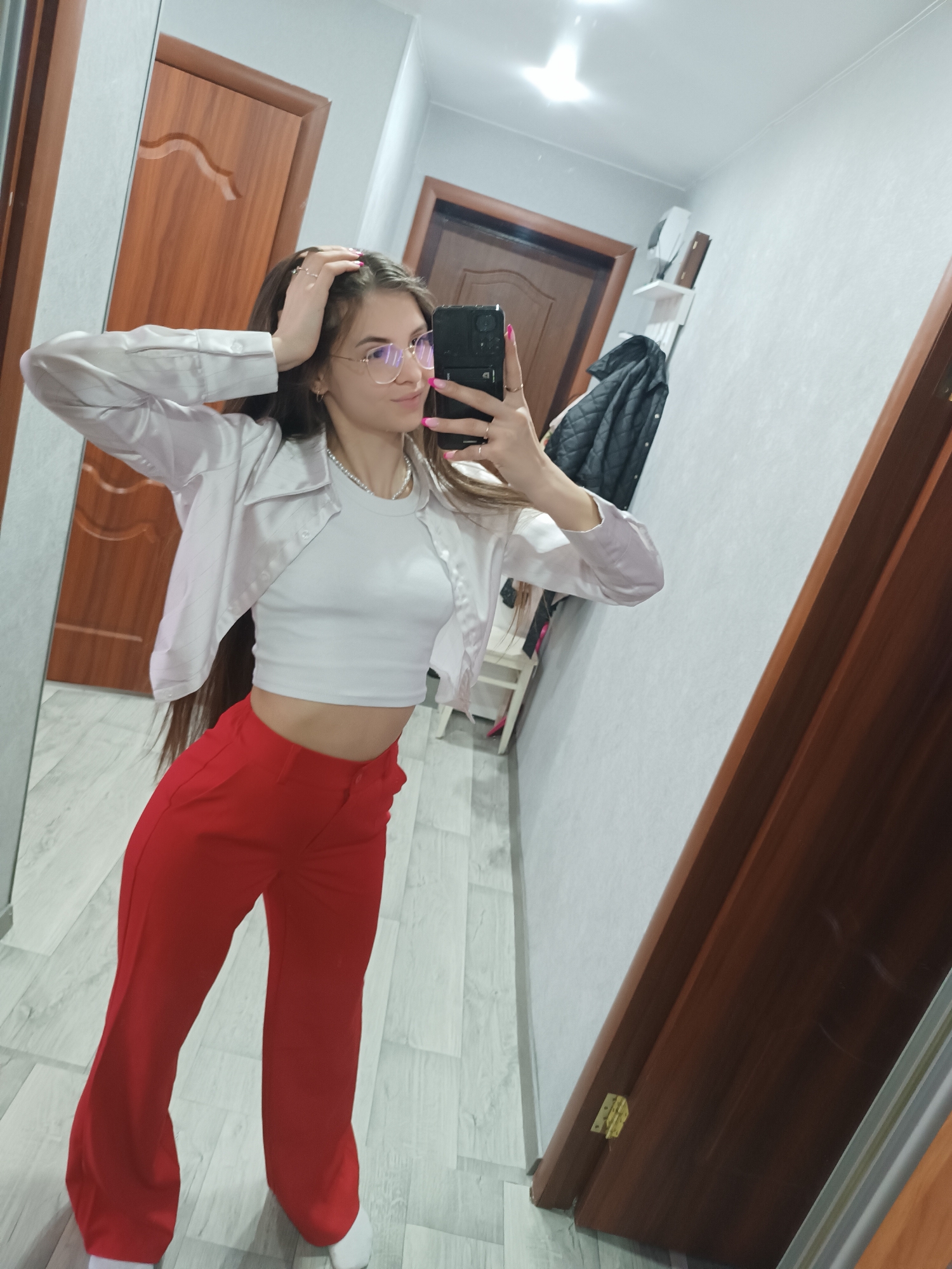 A young female subject is depicted taking a mirror selfie in an indoor setting, identified as a hallway or dressing area in the city of Unknown, Unknown. She stands facing a full-length mirror, holding a black smartphone with her right hand to capture the image, while her left hand is raised to her head, touching her long dark hair. The subject wears a white crop top, an open light pink/off-white collared shirt with long sleeves, red wide-leg trousers, and white athletic shoes. She is accessorized with gold-rimmed eyeglasses, a silver-colored beaded necklace, and multiple rings on her fingers, with light pink nail polish visible on both hands. The background, visible both directly and in reflection, features light grey textured walls, light wood-effect laminate flooring, and dark brown wooden doors. One door, reflected to the left of the subject, displays a curved panel design. To the right of the subject's reflection, a black quilted jacket hangs on a wall hook above a small white shelf. Overhead artificial lighting is visible in the mirror's reflection.