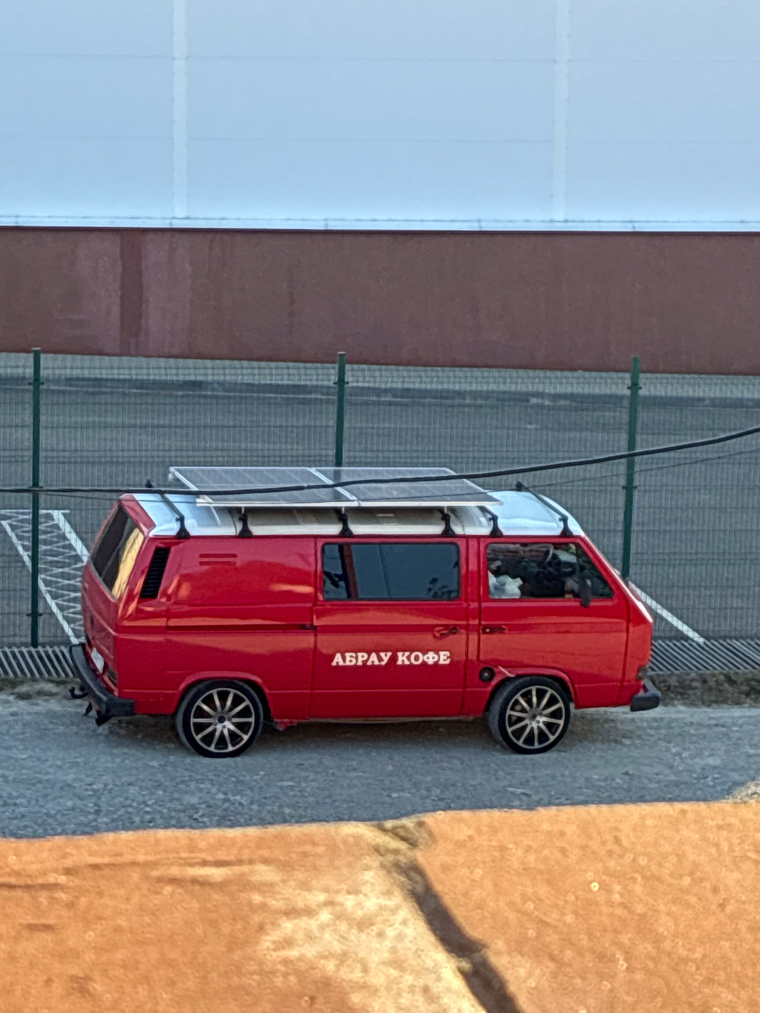 A red Volkswagen Type 2 van, with solar panels mounted on its roof rack, is parked on a paved surface. The van has the text "АБРАУ КОФЕ" (Abrau Coffee) in white lettering on its side. A green metal fence runs along the background, behind which is a building with a dark reddish-brown lower section and a light blue upper section. The ground in the foreground is a rough, orange-brown surface, possibly dirt or a textured path. The scene appears to be an outdoor area in the city of Abrau-Dyurso, Russia, given the inscription on the van. There is a person visible through the driver's side window of the van, appearing to be seated.
