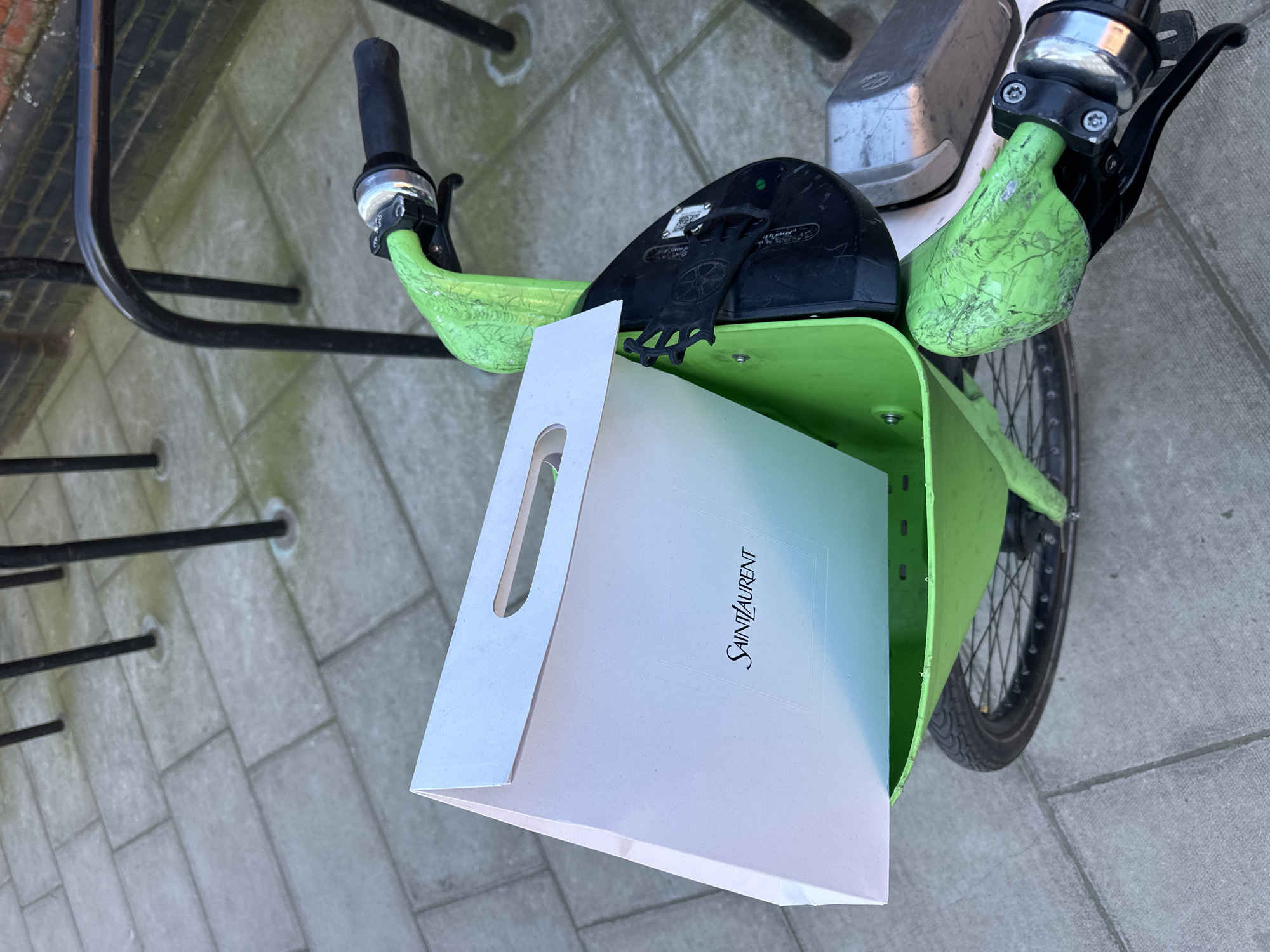 A white Saint Laurent shopping bag is positioned vertically within the bright green plastic front basket of a rental bicycle. The bag displays the "SAINT LAURENT" logo in black text and features die-cut handles. The bicycle itself is predominantly bright green, with black handlebars, grips, and brake levers. A black electronic control unit, featuring a QR code and a small green indicator light, is mounted on the front frame beneath the basket. The green paint on the handlebars exhibits visible wear and cracking. The bicycle is parked on a grey paved surface consisting of rectangular concrete pavers, with traces of moss visible in the joints and around the bases of black metal poles in the background. These poles, some forming a curved top, suggest a bicycle rack or urban barrier. A segment of a reddish-brown brick wall is visible in the upper right background. The scene is located in the city of London, United Kingdom. No individuals are present.