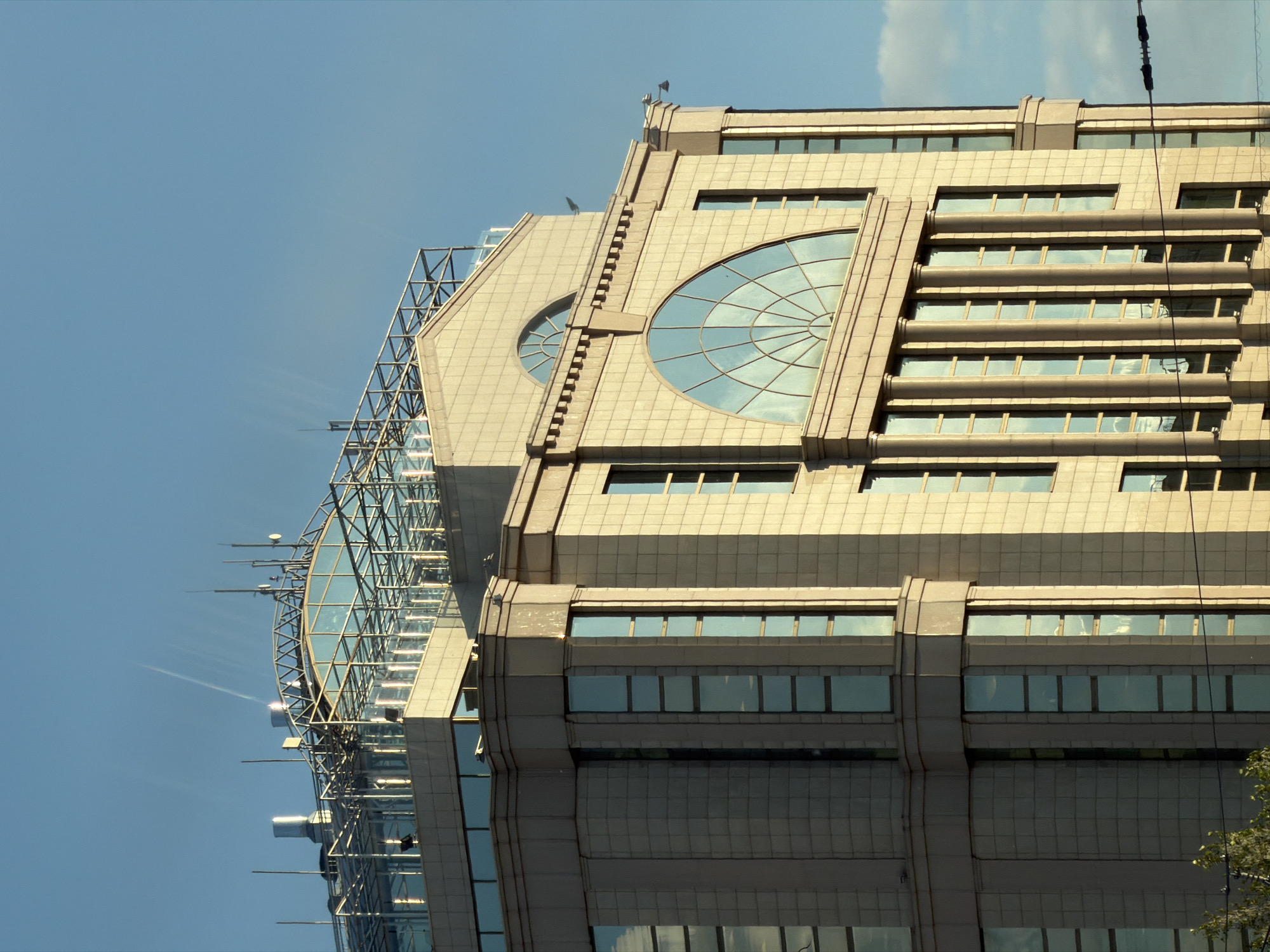 This media file presents a striking architectural close-up of a modern, multi-story building set against a clear, bright blue sky with a few wispy white clouds visible towards the top right. The building's facade is constructed from light-colored, possibly beige or cream-toned, stone or concrete panels, giving it a solid and imposing appearance.
A prominent feature of the building's design includes numerous dark, rectangular windows arranged in horizontal bands, reflecting the sky and ambient light. Two large, distinctive semi-circular windows with intricate radial spoke patterns are visible on one of the main faces, adding an artistic touch to the geometric structure. The upper left section of the building showcases a more contemporary style, featuring a complex glass and metal skeletal structure, possibly an enclosed atrium or rooftop garden, adorned with multiple antennae, poles, and other technological equipment.
The image is captured from a low angle, looking upwards, which accentuates the building's height and architectural details. Bright sunlight illuminates the structure, creating highlights on the panels and glass surfaces. The time of day appears to be sunny daytime, likely late morning or early afternoon. A dark cable or wire runs vertically down the right side of the frame, and a small amount of green foliage from a tree is visible in the bottom right corner. No people or explicit activities are depicted, focusing entirely on the building's static presence. There is no visible text in the image. The location context for this building is Moscow, Russia.