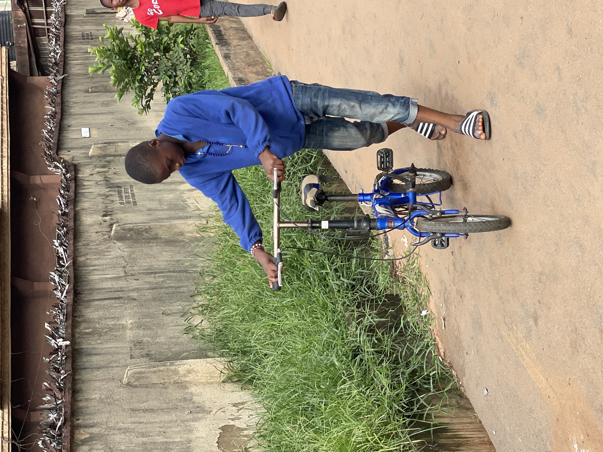 A young male wearing a blue sweatshirt and jean shorts stands with one foot on a blue bicycle. He holds an object in his left hand. He is wearing Adidas slides. A boy in a red shirt stands behind him. The location appears to be on a sidewalk with a concrete wall to the left and grass on the ground. This is occurring in Onitsha, Nigeria.