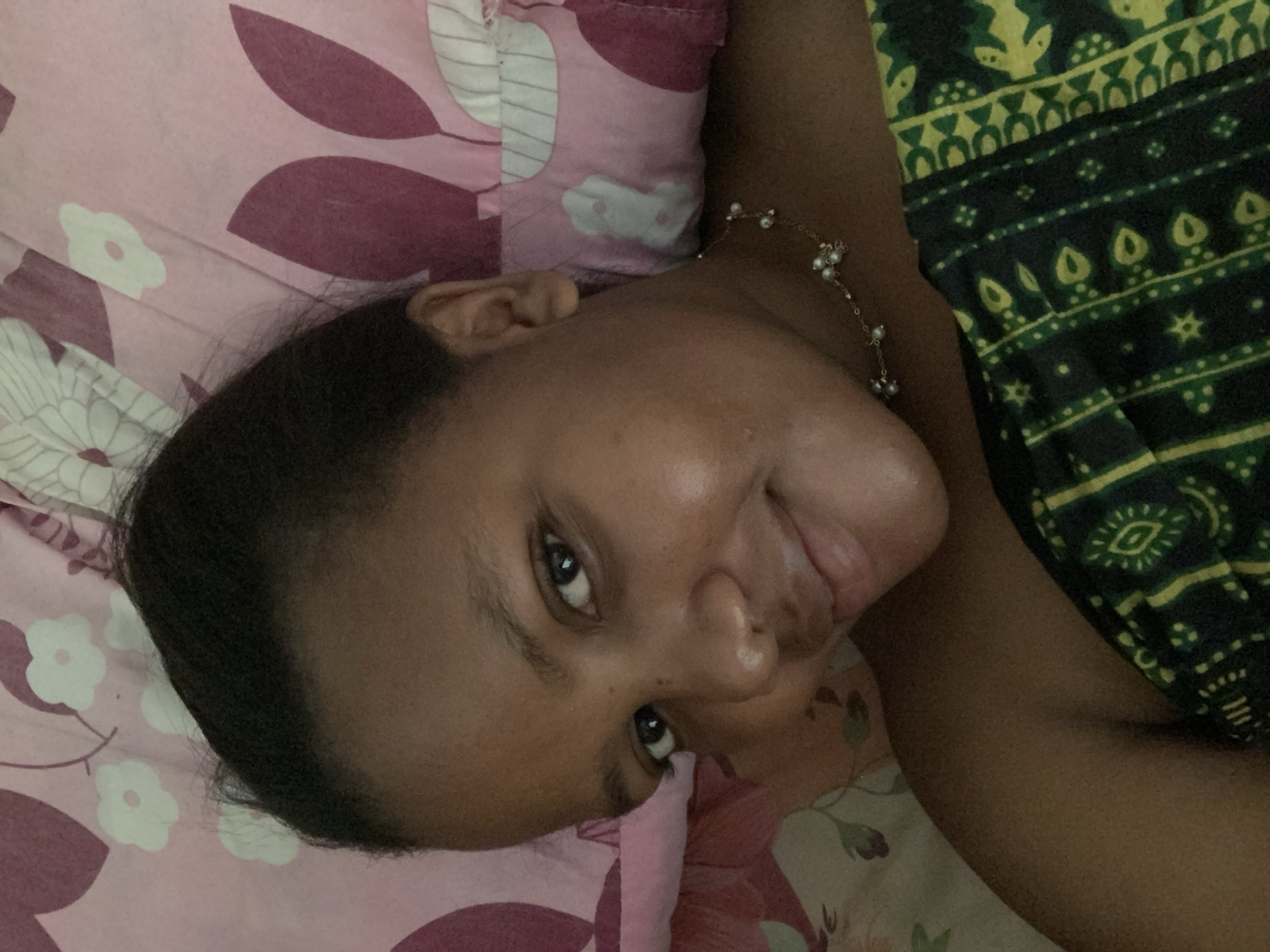 A young woman with dark hair and brown skin lies on her side on a bed with floral patterned bedding. She wears a delicate necklace with small pearls. Her head rests on a pink floral pillow. She is partly covered by a dark green and yellow patterned garment. Her face is turned towards the camera, with a slight smile. It is not possible to ascertain the location within Dar es-Salaam, Tanzania.