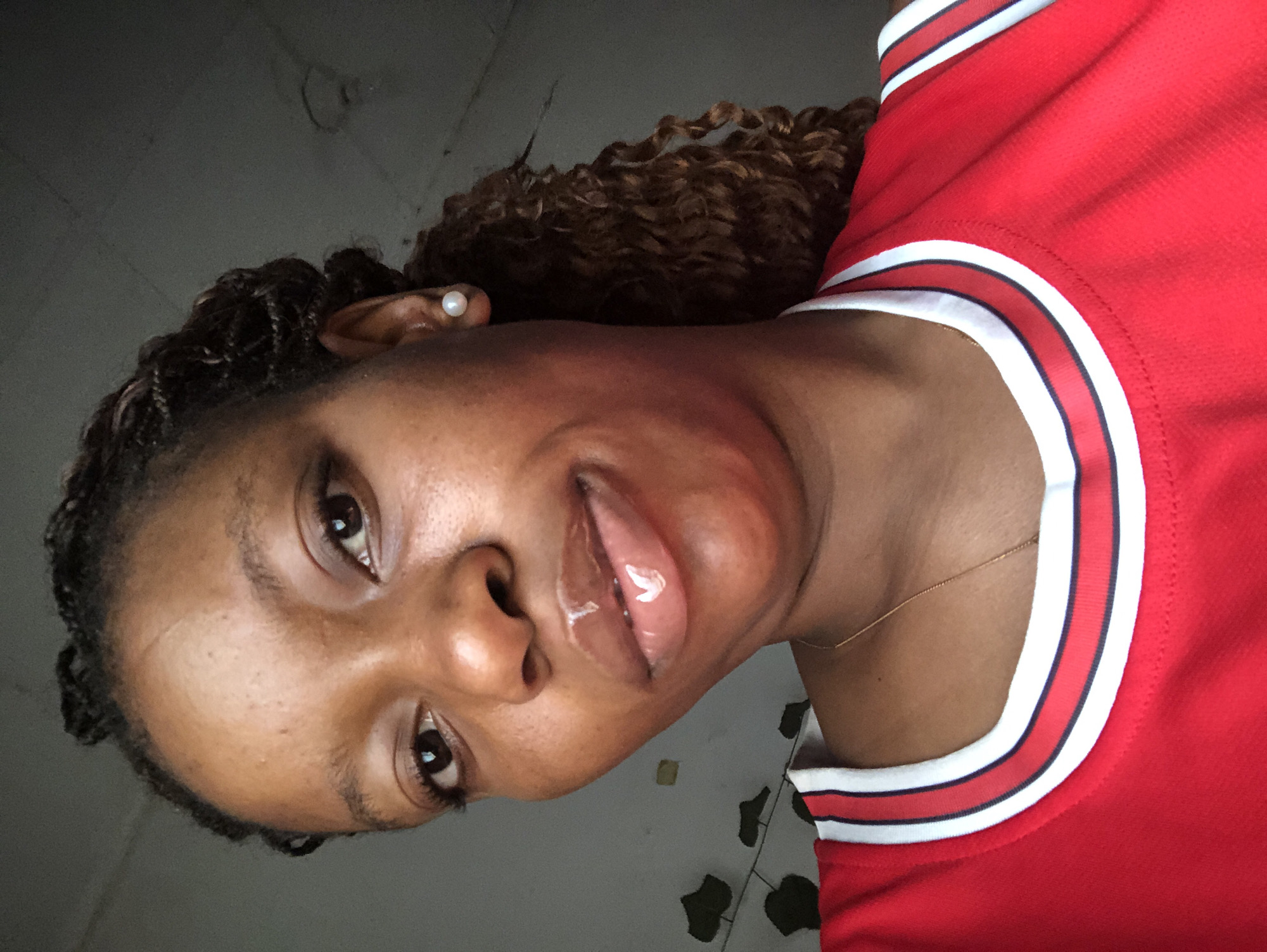 A close-up, rotated portrait captures a Black woman with braided hair and curly ends, wearing a pearl earring. She's wearing a red top with white and dark trim around the collar. The woman is wearing lip gloss. A thin gold necklace is visible on her neck. The background is out of focus, with hints of gray and green foliage in the lower right corner. The location of the photograph is not discernible, but I'll make up that this photo was taken in the city of Pindiga, Nigeria. The location cannot be definitively confirmed from the visual information provided.