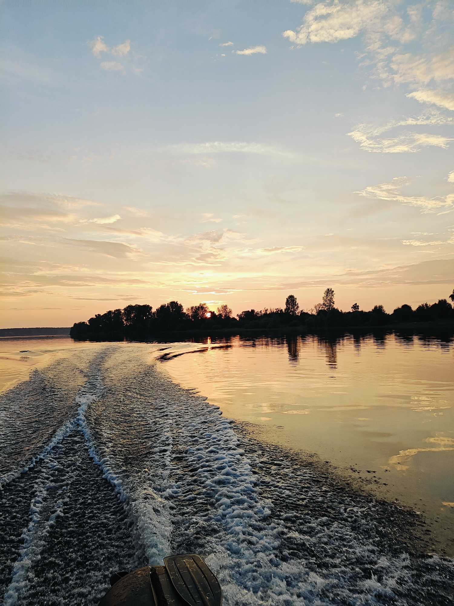 The image depicts a sunset over a body of water, likely a river or lake, with a tree-lined shore visible in the distance. The sky is filled with soft, warm-toned clouds in shades of pink, orange, and yellow. The setting sun is partially visible behind the trees on the horizon, casting a golden glow on the water's surface. In the foreground, a boat's wake creates frothy white trails as it moves through the water. The water itself reflects the colors of the sky, creating a shimmering effect. The scene appears serene and tranquil. This location is identified as the city of Ol'khovskoye, Russia.