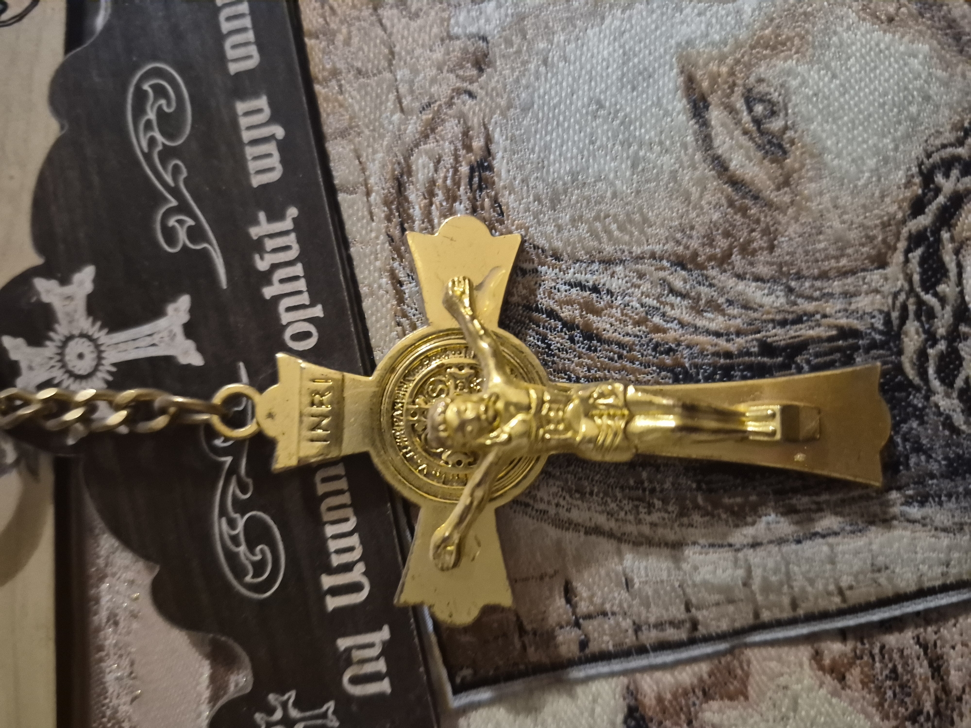 A close-up, eye-level shot shows a gold-colored crucifix hanging from a chain. The crucifix depicts Jesus on the cross and bears the inscription "INRI" at the top. The crucifix is positioned in front of a dark-colored ornamental plaque with white etchings of crosses and decorative swirls. Below the plaque and crucifix is a textured tapestry or fabric with an image of a person's face, particularly focusing on the eye and hair. The scene is likely indoors, possibly in a home or religious setting in Yerevan, Armenia, given the Armenian script visible on the plaque.