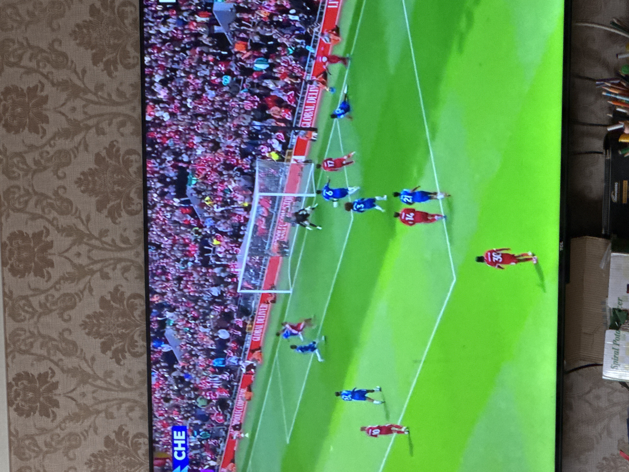 A television displays a soccer match. The pitch is green, with players in blue and red uniforms. The crowd is visible, densely packed and wearing red clothing. The game appears to be near a goal. The television is indoors, set against a backdrop of patterned wallpaper and miscellaneous items. The image was not taken in Yerevan, Armenia.