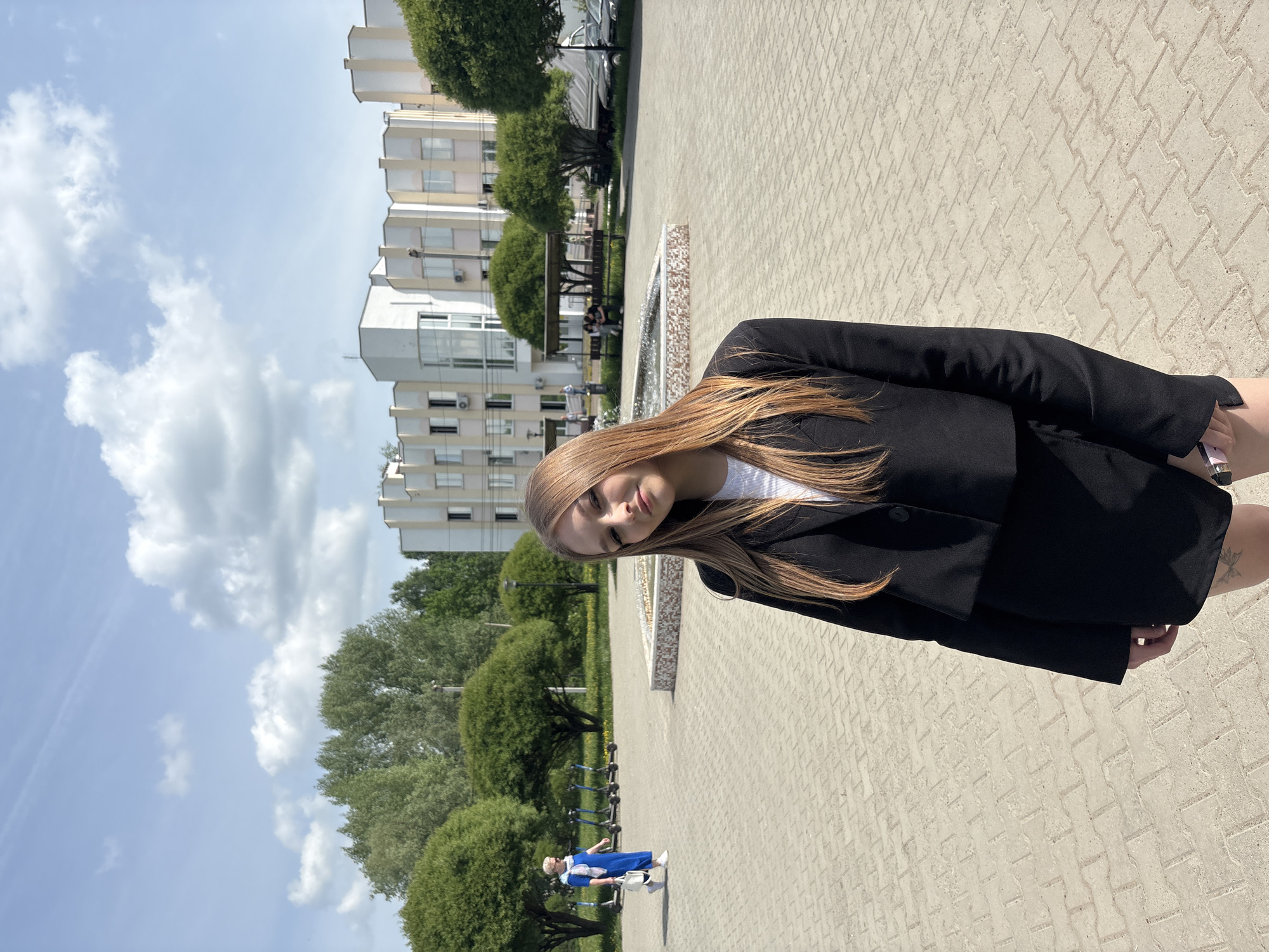 A young woman with long, light brown hair stands on a paved walkway in Chaikovsky, Russia, next to a fountain. She is wearing a white shirt and a black blazer. In the background are apartment buildings, trees, benches, and a cloudy sky. A person dressed in blue is pushing a shopping cart along the same walkway. The image is a daytime shot with bright sunlight.