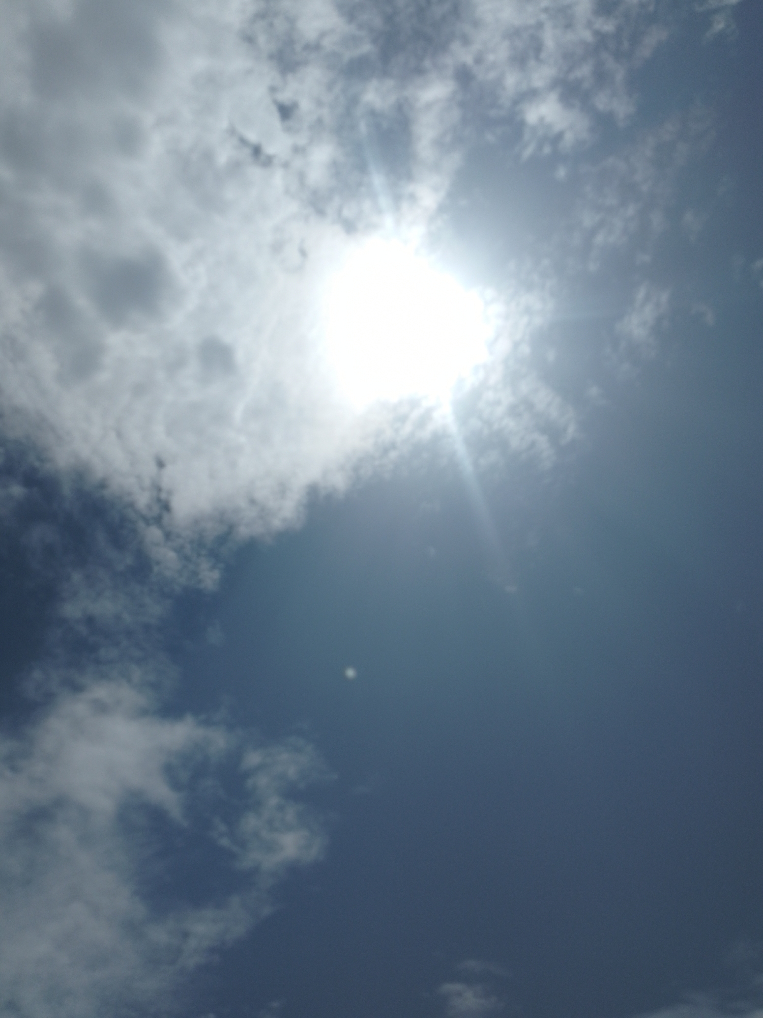 The image shows a sky filled with clouds. The sun is shining brightly and is partially obscured by clouds, creating a lens flare effect. The sky is a deep blue, suggesting it is daytime, likely late morning or early afternoon. There are scattered white clouds and darker, grayer clouds, indicating a partly cloudy or overcast sky. In the distance, there appears to be a small, bright object in the sky, possibly a distant cloud or a lens artifact. The image is taken from a low angle, looking up at the sky. There is no text visible in the image. The overall mood is bright and airy, characteristic of a sunny day, though the presence of darker clouds hints at changing weather. The scene is natural and vast, focusing on the elements of the sky.