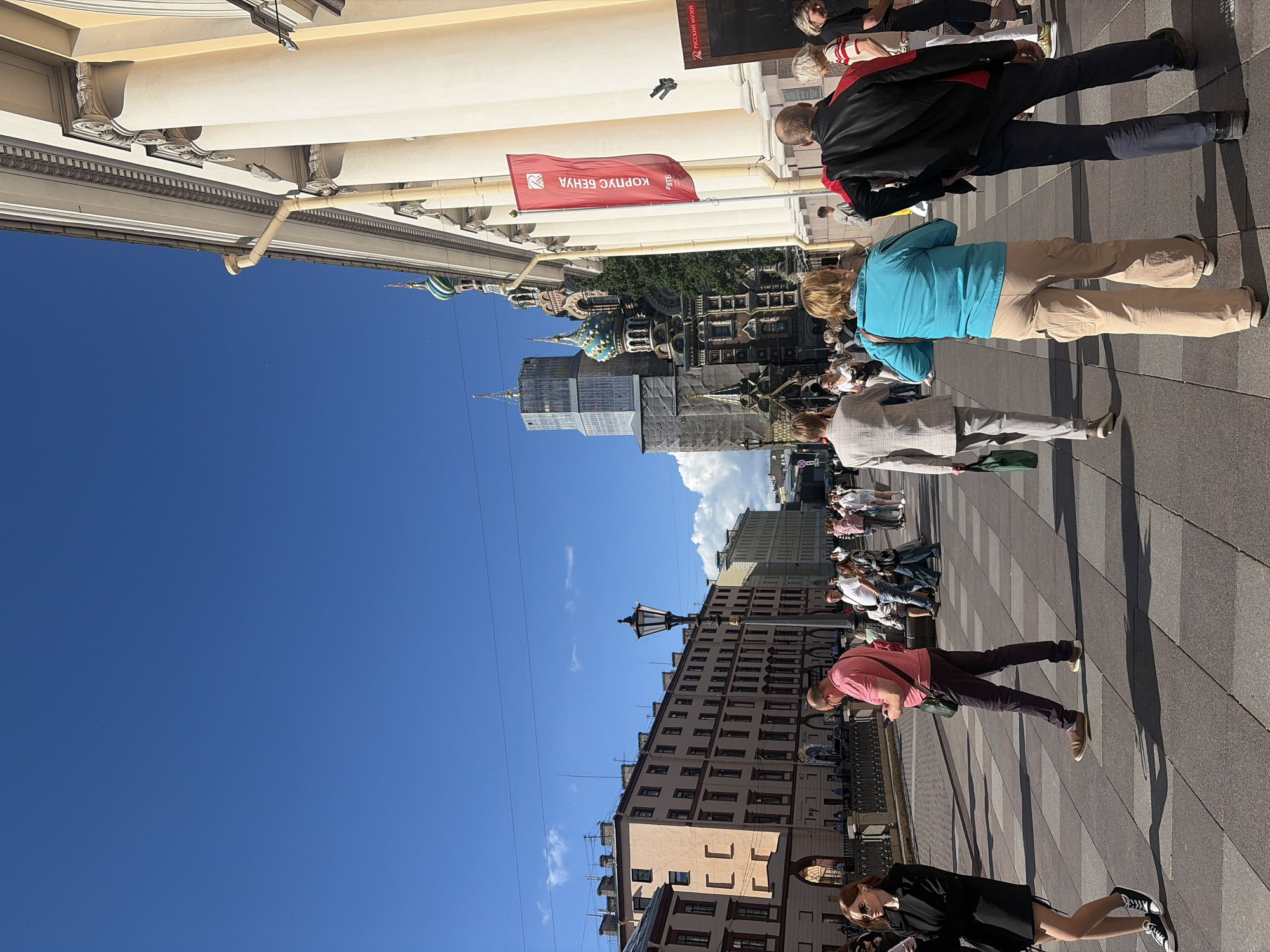 The image captures a street scene in Saint Petersburg, Russia, on a clear, sunny day. The perspective is tilted, making it appear as if the viewers are looking up from a street level towards a bright blue sky. Several people are visible walking along a paved sidewalk, casting distinct shadows due to the strong sunlight, suggesting it is midday.
In the background, a mix of architectural styles is evident. A grand, light-colored building with ornate columns lines the upper part of the frame, suggesting historical architecture. Further back, a cluster of distinctive onion-domed churches, possibly part of a cathedral complex like Saint Basil's or Kazan Cathedral, adds to the iconic Saint Petersburg skyline. A modern skyscraper is also visible, juxtaposing the historic and contemporary elements of the city.
One person, wearing a blue jacket and beige pants, walks towards the viewer. Another individual in a black jacket and dark pants is also present. A red flag with white Cyrillic text is prominently displayed, reading "КОРПУС БЕНУА" (Corpus Benois). This likely refers to the Benois Wing of the Russian Museum. The overall atmosphere appears to be one of urban activity and sightseeing, with people enjoying the pleasant weather.
