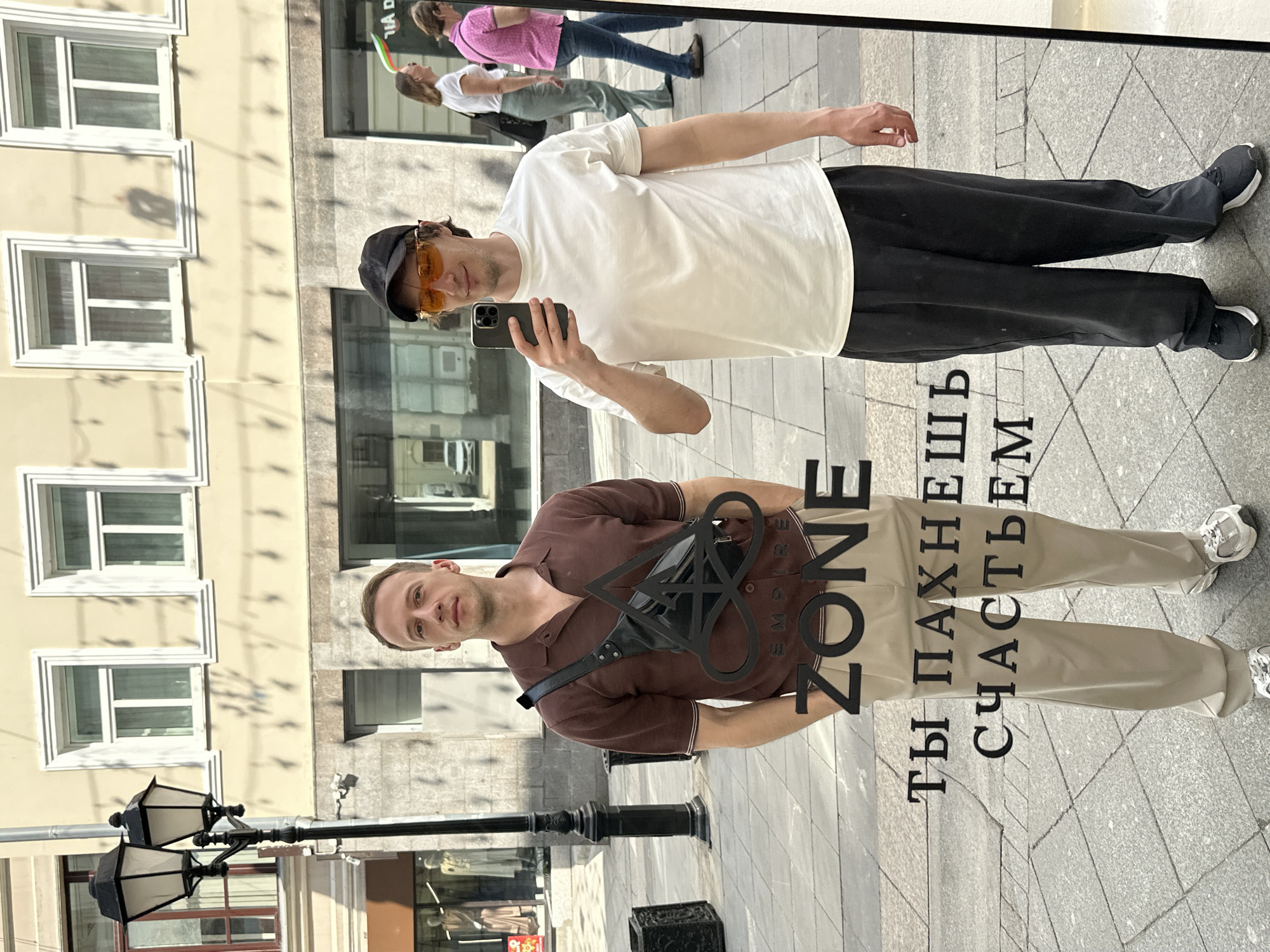 The image shows two people reflected in a mirror, likely taking a selfie in an urban environment. The mirror is located in Moscow, Russia, as evidenced by the Russian text "ТЫ ПАХНЕШЬ СЧАСТЬЕМ" which translates to "YOU SMELL LIKE HAPPINESS".
The upper reflection shows a man wearing a white t-shirt, black pants, a black cap, and orange-tinted glasses. He is holding a smartphone and appears to be taking a picture. In the background, other people can be seen walking.
The lower reflection shows a man wearing a brown polo shirt, light-colored pants, and a black fanny pack. The logo "ZONE EMPIRE" is superimposed in the mirror.
The background of the scene shows a building facade with windows and street lamps. The street is paved with light gray stones.