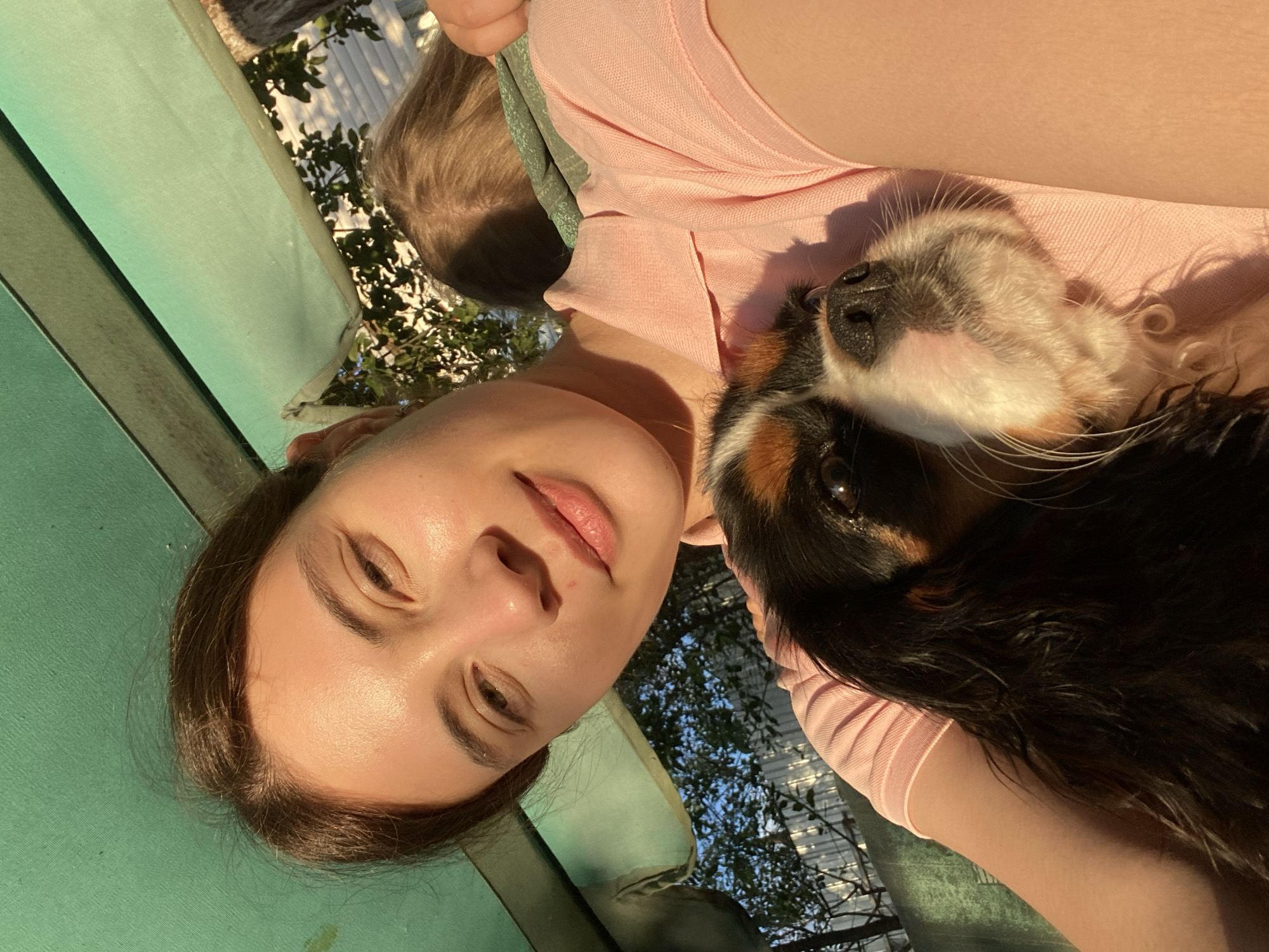 A young woman with dark hair is holding a Cavalier King Charles Spaniel in her arms. She is wearing a pink polo shirt. The dog's head rests on her shoulder, and its eyes are looking towards the camera. The background is out of focus, but appears to be an outdoor setting with greenery and a fence. The lighting suggests it is daytime, with sunlight casting shadows. The overall impression is one of a casual, affectionate moment. There is no information available to confirm the location in Votkinsk, Russia.