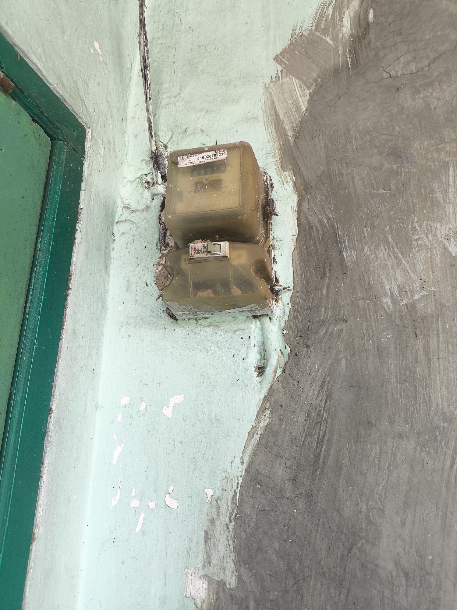 A close-up shot captures an outdoor wall in Lumajang, Indonesia, featuring a green door frame on the left and an electricity meter mounted on a light green, weathered wall. The meter is enclosed in a translucent plastic casing, with a digital display visible and a switch below it. The wall shows signs of peeling paint and repairs, with a section of rough, gray plaster encroaching from the right side of the frame. A black electrical cable runs vertically along the wall, disappearing into the top of the meter. The overall impression is one of a functional, yet aged and somewhat neglected, exterior utility installation.