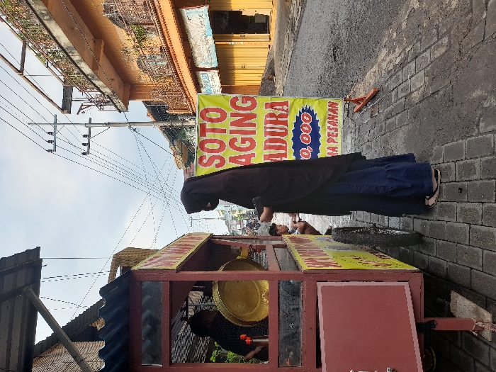 A street food vendor is pushing a cart with a sign reading "SOTO AGING MADURA 10,000." A person in a dark hijab and blue dress stands beside the cart on a paved street. The cart is topped with a yellow surface and has a maroon side. A golden bowl is visible inside the cart. Buildings and utility poles line the background.