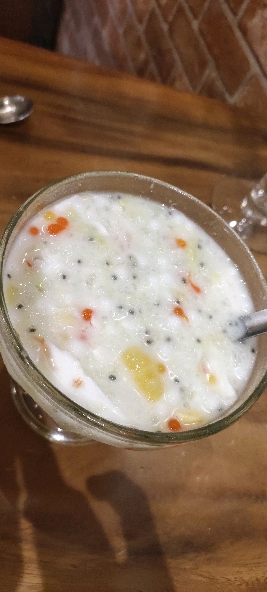 An overhead shot shows a clear glass bowl filled with a white, creamy dessert. The dessert is topped with small, black seeds and pieces of orange and yellow fruit. The bowl sits on a wooden table. To the left of the bowl, a small silver spoon is visible. In the background, a wall with exposed brick is visible. The lighting suggests an indoor setting. The image is focused on the dessert, with the background slightly blurred. There are no people visible in the image.