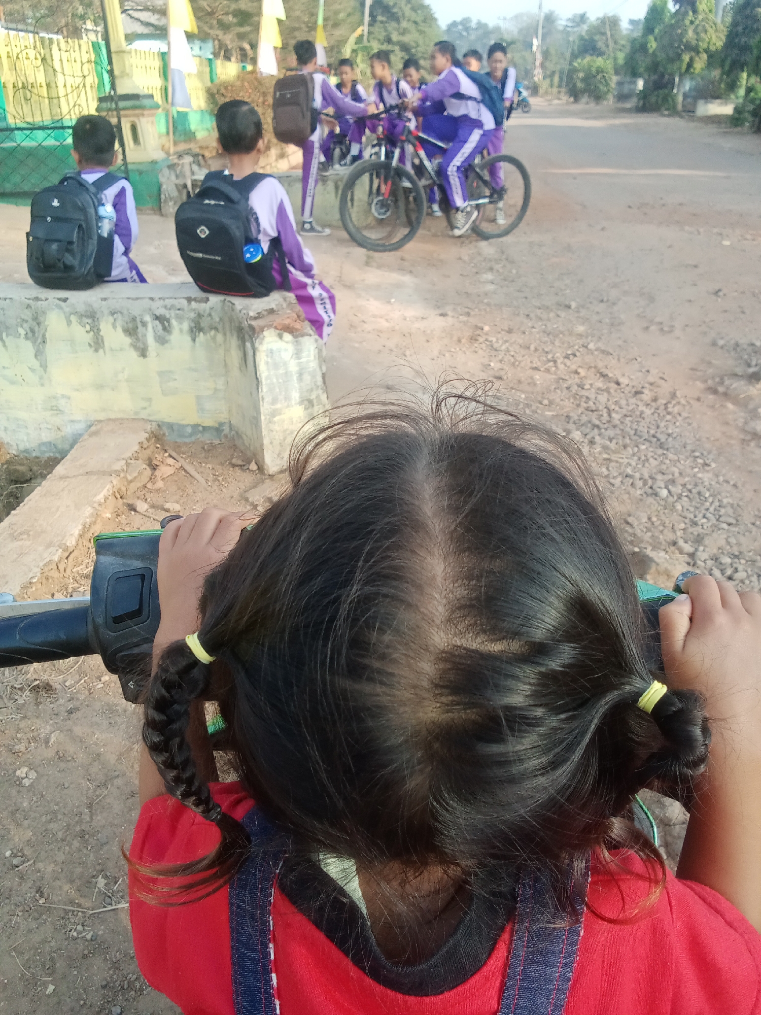 In Sri Sawahan, Indonesia, two young children, one in the foreground and another partially visible, are seated on a concrete ledge. The child in the foreground, viewed from behind, has dark hair styled in two braids secured with yellow elastics. They wear a red shirt with denim overalls and hold onto the handlebars of a vehicle, possibly a scooter or motorcycle. Behind them, another child, also viewed from behind, has a black backpack and wears a purple and white uniform.
In the background, a group of children in similar purple and white uniforms are gathered near bicycles. Some of them are interacting with the bicycles, and one appears to be riding one. The environment is outdoors, with a dirt road and scattered gravel. Vegetation, including trees, is visible in the distance, and some structures, possibly part of a school or community area, are present. The lighting suggests it is daytime, possibly morning or late afternoon, with soft sunlight.