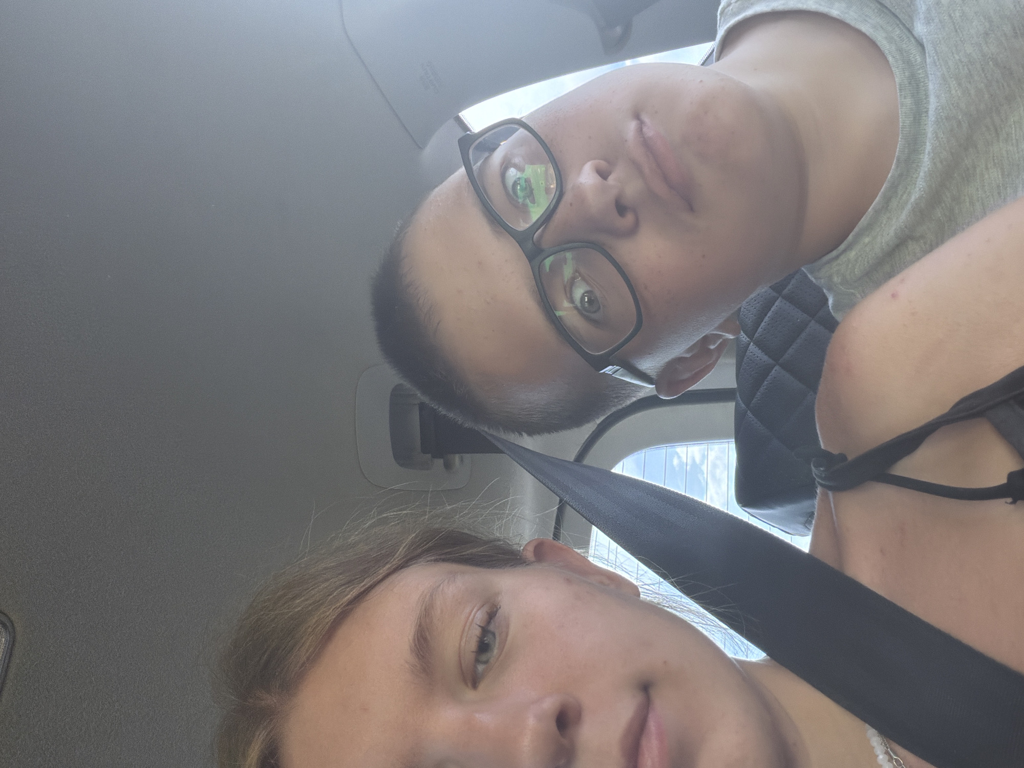 Two young people, a girl and a boy, are in the backseat of a car. The girl is on the left, with blonde hair and a seatbelt across her chest. She is looking forward with her mouth slightly open. The boy is on the right, wearing glasses and a grey t-shirt. He is looking towards the camera with a neutral expression. The background shows the interior of the car, including a headrest and a window with a view of the sky. The image appears to be taken in Novoe Feizullovo, Russia.