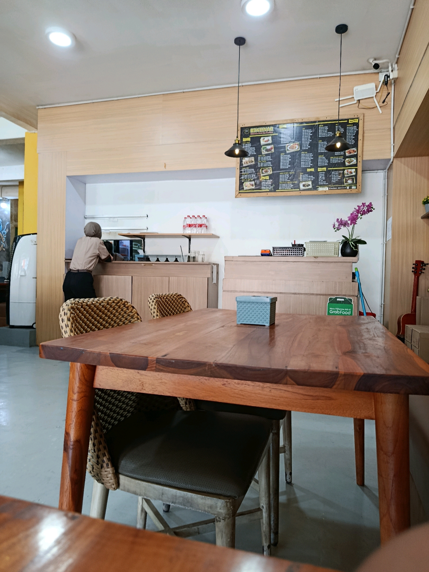 This image depicts the interior of a restaurant in Bekasi, Indonesia. In the foreground, a rustic wooden dining table is the primary subject, with two chairs visible beneath it. One chair is made of woven material and wood, while the other appears to be upholstered in dark fabric with a wooden frame. A small, light blue rectangular container sits on the table. In the background, a woman wearing a headscarf and a light brown uniform is seen standing behind a counter, facing away from the camera. She appears to be interacting with equipment or items on the counter.
Behind the counter, a menu board is prominently displayed on the wall. The menu board is black with white text and features images of food items. Two pendant lights hang from the ceiling, illuminating the area. To the right of the menu board, a security camera is mounted on the wall. Further to the right, a red acoustic guitar leans against the wall, and some cardboard boxes are stacked nearby. A potted orchid with purple flowers adds a decorative touch to the counter area. On the counter itself, there are several bottles of water, cups, and other small items. The environment appears to be a clean and functional dining space.