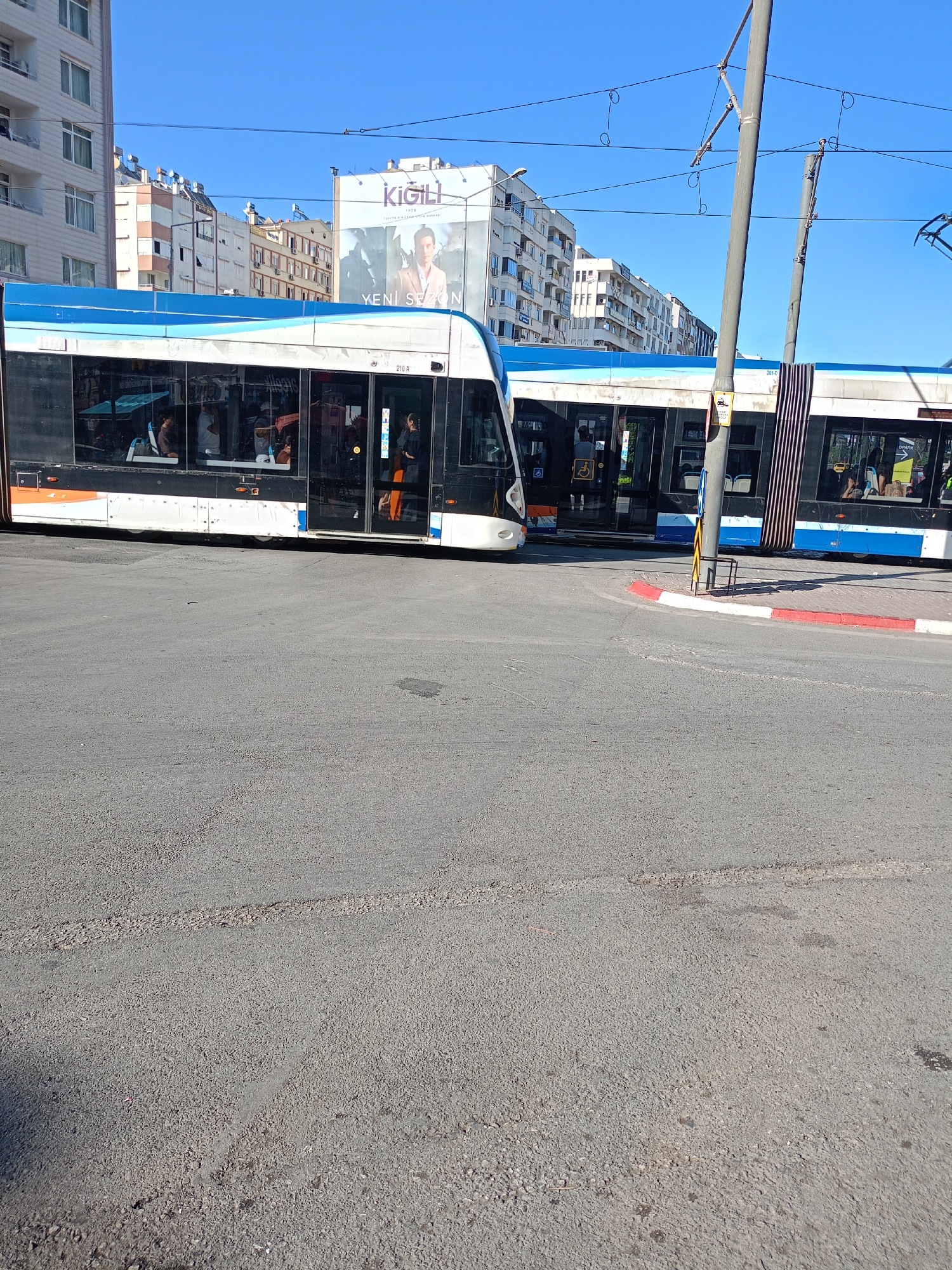 A bright, sunny daytime exterior shot captures a modern tram on a street corner in Antalya, Turkey. The tram, predominantly white with blue accents and dark tinted windows, is a key subject. Several passengers are visible inside, including a person standing in the doorway, a person in an orange garment, and others seated. The tram is positioned at an intersection with a red and white curb.
Behind the tram, several multi-story buildings of varying architectural styles line the street. One building features a large billboard with an image of a man and text that reads "KIGILI YENI SEZON". Another building has a prominent blue and white wavy design along its upper floors. Overhead, a network of electrical wires for the tram system crisscrosses the clear blue sky. The street is paved with asphalt, and there are visible tire tracks and some minor debris on the surface. A metal pole, likely part of the tram's electrical infrastructure, stands near the intersection. The overall scene depicts urban transportation and commerce in a Turkish city.
