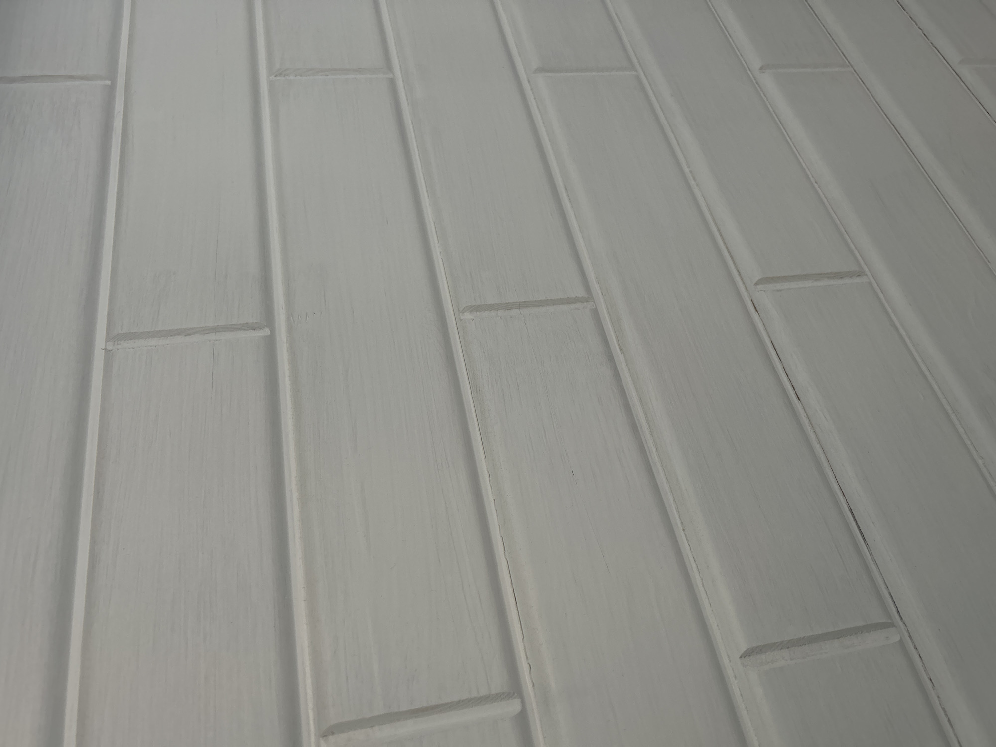 The image shows a close-up, angled view of a light gray, textured wall made of horizontal rectangular panels that resemble painted wood planks or large tiles. The panels are arranged in a staggered pattern, with visible vertical and horizontal grout lines or seams between them. The surface of the panels has a slightly distressed or brushed appearance, with visible grain-like textures and subtle variations in color, suggesting it is painted or finished wood. The lighting casts soft shadows, emphasizing the slight depth and texture of the panels and their edges. The scene is devoid of people, animals, or other objects, focusing solely on the architectural detail of the wall. The location is unknown.