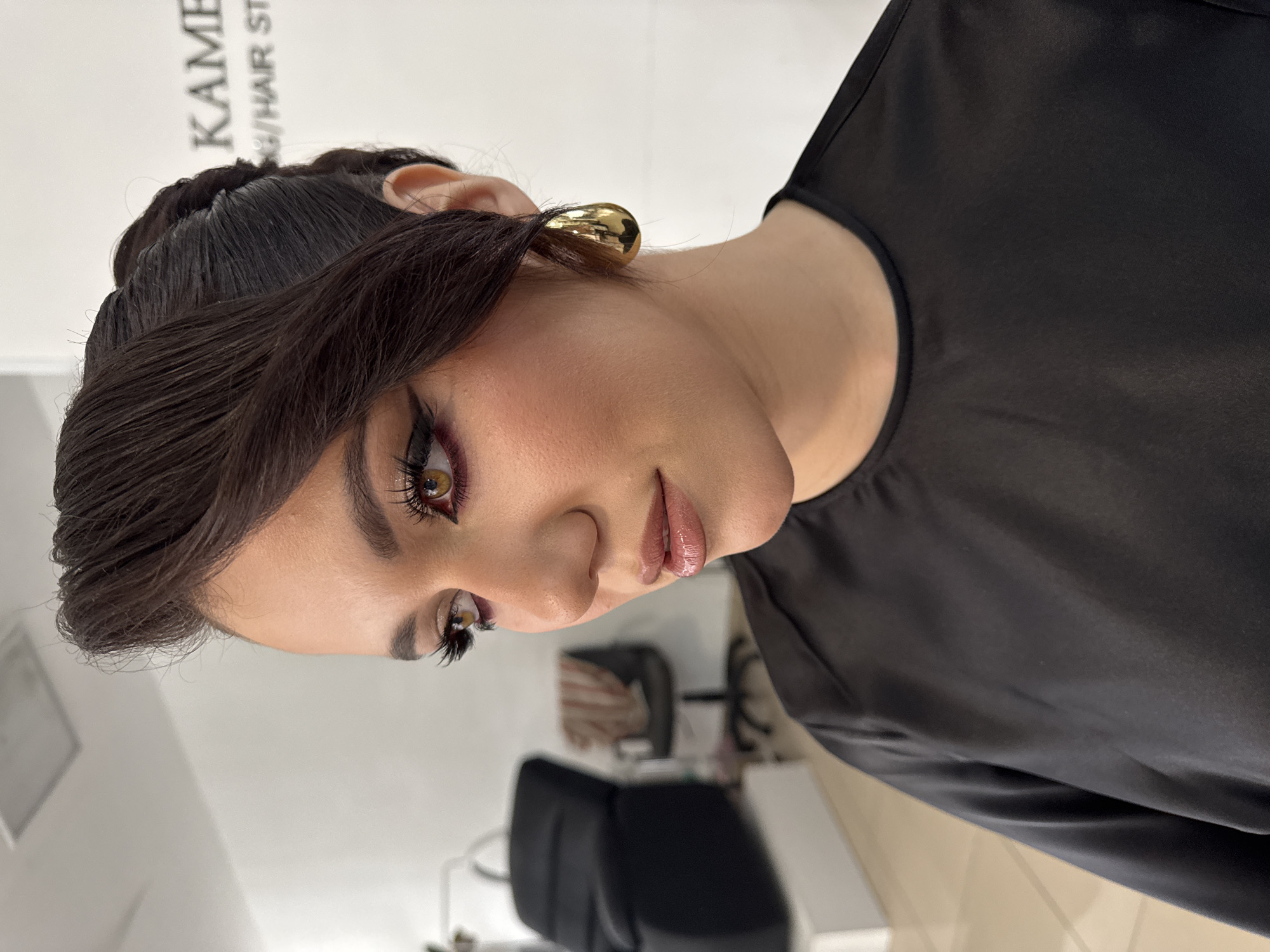A portrait of a woman is captured indoors. The woman has dark hair styled off her face and is wearing gold earrings. She has full makeup, including eyeliner and lip gloss. She is wearing a black top. The setting appears to be in a salon or beauty studio. The photo was taken in the city of Chaikovsky, Russia. A chair and some blurry equipment are visible in the background.