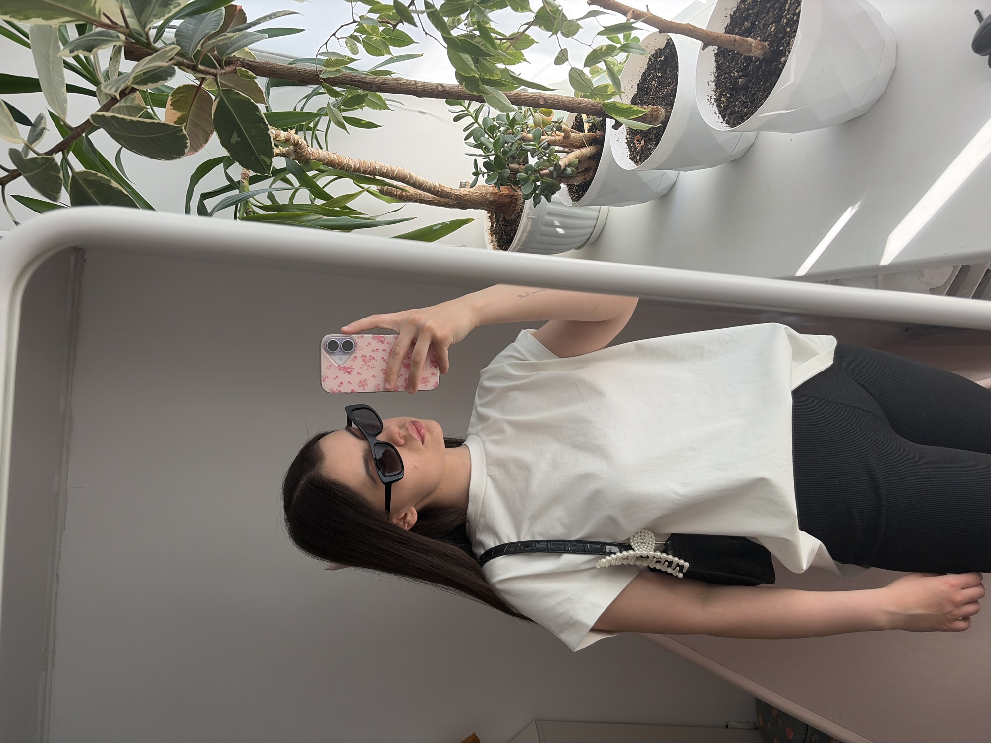 A woman is taking a selfie in a mirror in Chaikovsky, Russia. She is wearing sunglasses, a white shirt, and black pants. She is holding a pink phone with a floral pattern. There are several plants in white pots visible in the background. The background includes a wall.