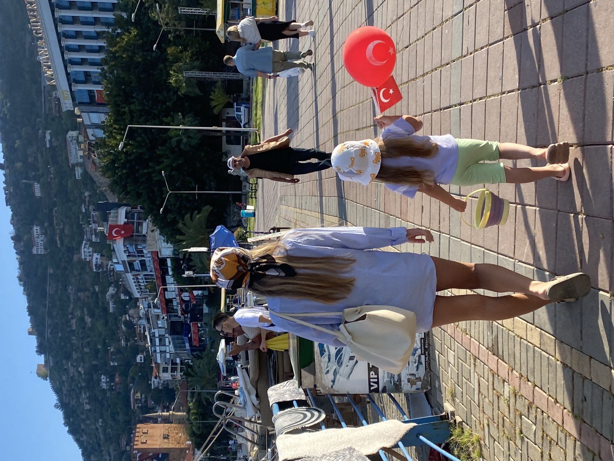 This outdoor image, taken on a clear, sunny day in Alanya, Turkey, depicts a vibrant scene along a paved promenade or harbor walk. The perspective is from slightly behind and to the left of several people walking.
In the foreground, a woman with long, light brown hair, dressed in a light blue long-sleeved tunic or dress and a patterned headscarf (orange, black, and white), walks away from the viewer. She carries a cream-colored shoulder bag. To her right, a young girl with blonde hair, wearing a light blue top, light green shorts, and a white sun hat with dog motifs, also walks. She holds a red balloon featuring the Turkish flag emblem (white crescent moon and star) in her right hand and a small Turkish flag on a stick in her left, while carrying a yellow and purple striped bucket.
Further along the path, a man in a light brown jacket, dark pants, and a white hat is visible. Behind him, other individuals, including a woman in a black skirt and a man in a light blue shirt, are present. To the left, near what appears to be a railing overlooking the water, a man in sunglasses and a white t-shirt looks at his phone.
The setting is characterized by a paved walkway with a brick pattern. To the left, boats are docked in a marina or harbor. Across the water and nestled into verdant, tree-covered hills, a town or city rises. A large building on the hillside prominently displays the text "KAPTAN & GÜLVERT." Another Turkish flag is visible on a building higher up the hill. The strong shadows cast by the people indicate bright sunlight, suggesting midday or early afternoon. The overall atmosphere is relaxed and pleasant, with the Turkish flags adding a national or celebratory touch.