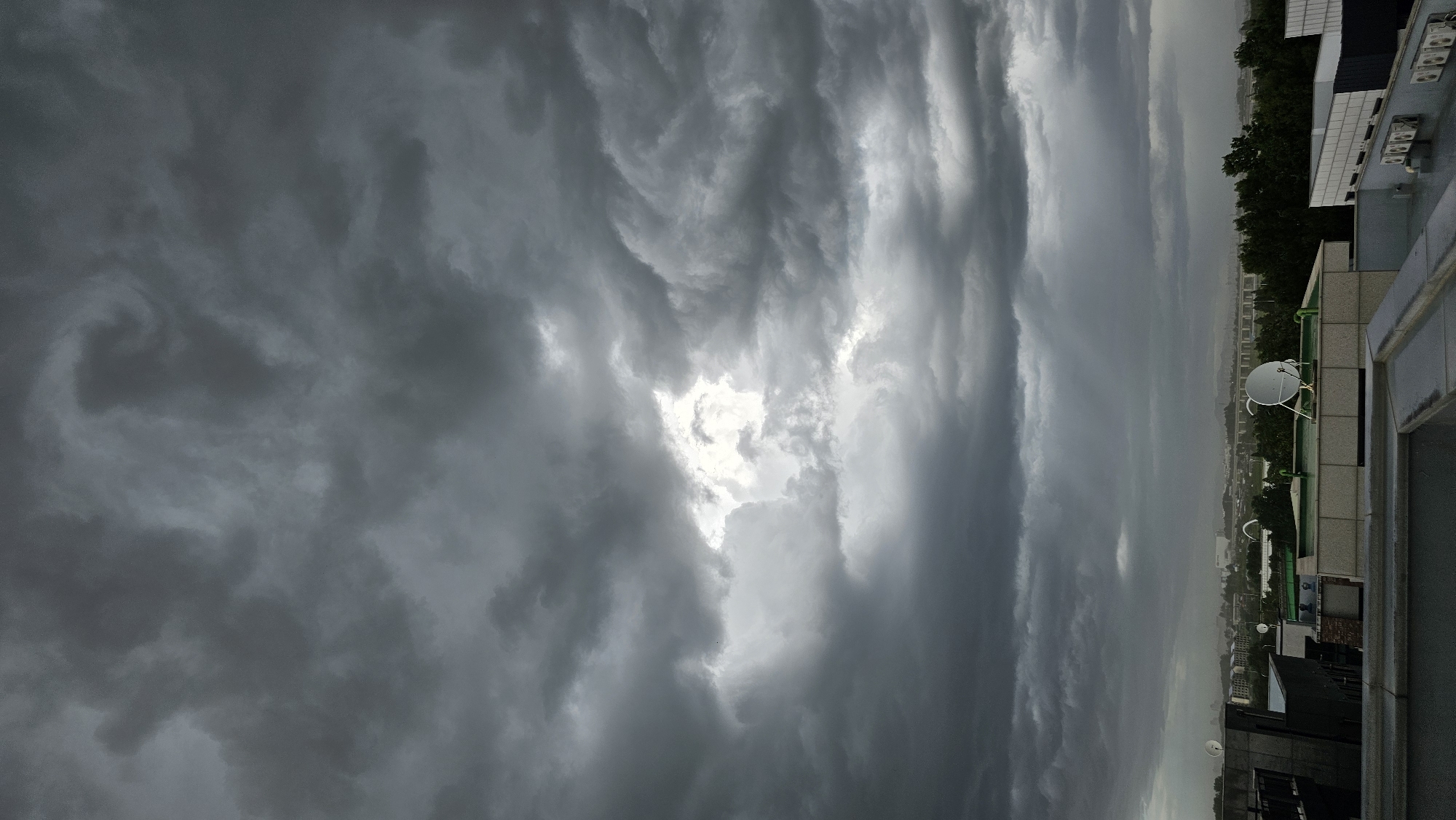 A vertical, high-angle shot captures a brooding sky over Asan-si, South Korea, just before or during a rain shower. Dark, heavy clouds dominate the frame, with a patch of brighter sky peeking through the center. Below the ominous clouds, a cityscape unfolds, featuring a variety of buildings, including some with satellite dishes and air conditioning units visible on their rooftops. A dense line of dark green trees separates the urban foreground from a more distant, hazy landscape. The overall impression is one of impending or ongoing inclement weather.