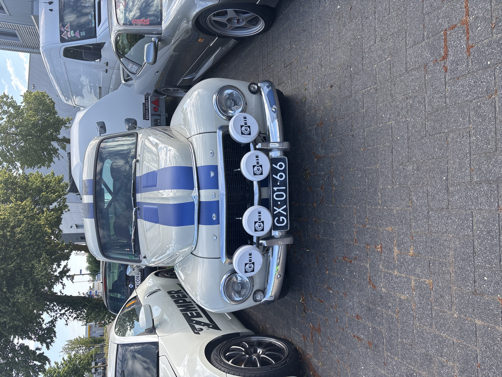 The image shows the front of a vintage white and blue Volvo PV544 parked on a paved surface. The car has two Cibie fog lights mounted on its front bumper and a Dutch license plate that reads "GX-01-66". To the left of the Volvo is a white Nissan 350Z, and to the right is a silver Nissan Silvia S15. Behind the Volvo is a white trailer. The scene appears to be outdoors, possibly a parking lot, with trees and buildings visible in the background. There is no indication of the specific location in Asten, Netherlands.