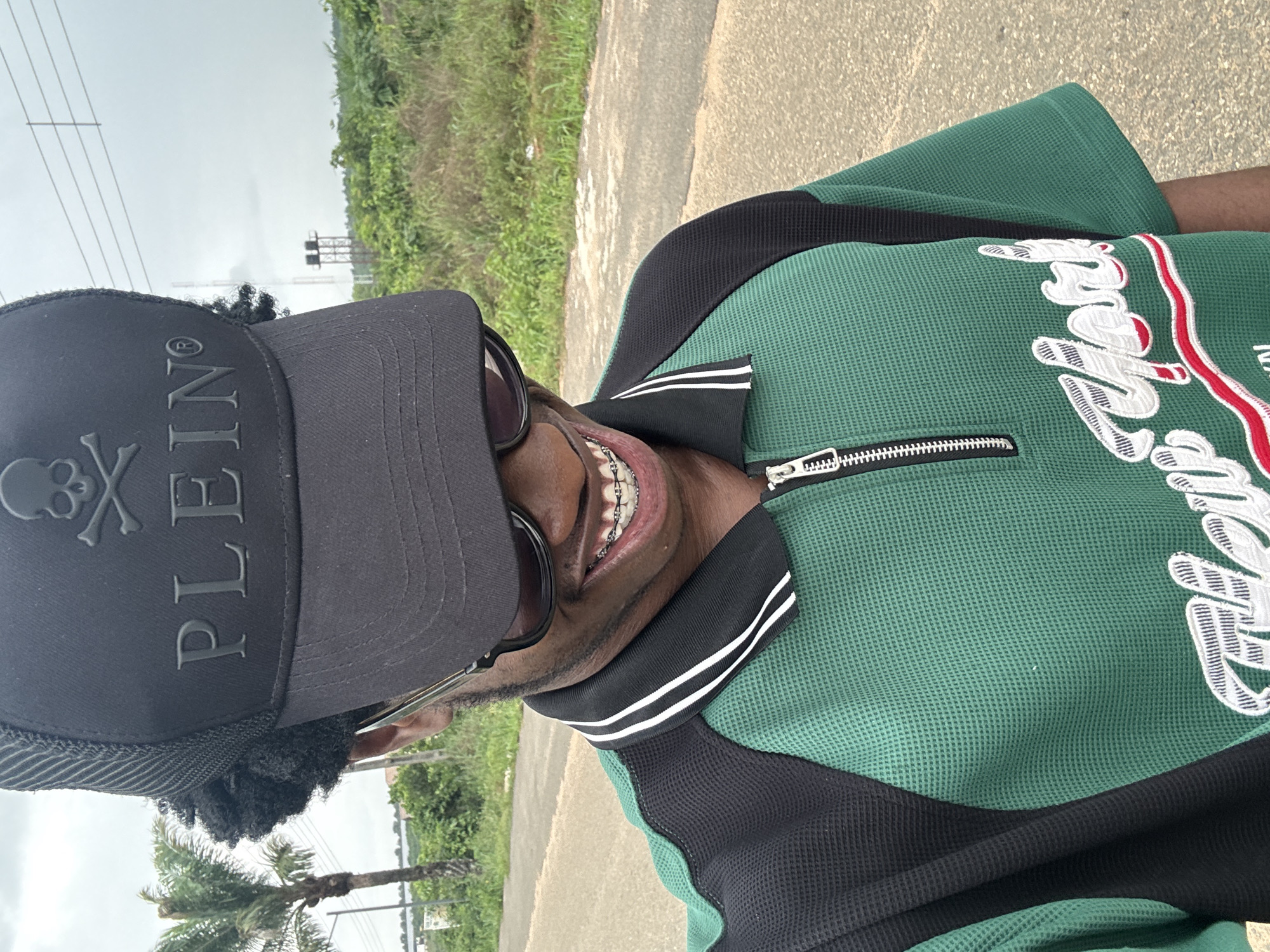 A close-up shot of a man wearing a black PLEIN baseball cap with a skull and crossbones logo, sunglasses, and braces. He's wearing a green and black zippered shirt with white and red embroidered lettering on the chest. He is standing on a paved area, possibly a sidewalk or road, with green vegetation and trees in the background. Overhead power lines are visible. Given the prompt, the location is implied to be in Ihiagwa, Nigeria.