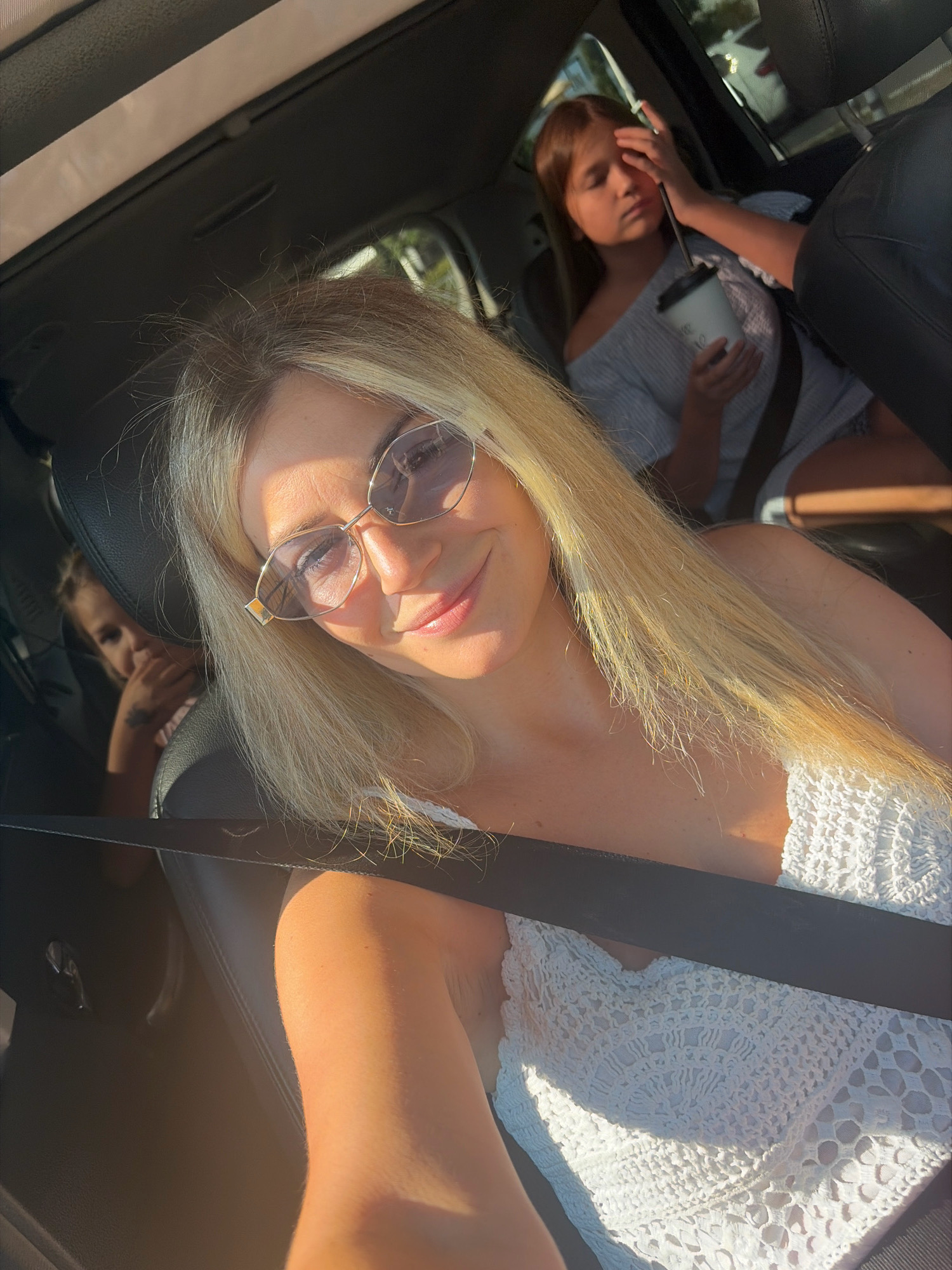 In a sunlit vehicle, a woman with blonde hair and sunglasses is taking a selfie, smiling. She is wearing a white crocheted top and a seatbelt. In the background, two children are seated in the back. The child on the right, with long brown hair, is holding a cup and touching her face. The child on the left is partially visible, with their hand near their face. The vehicle appears to be in motion, with light filtering through the sunroof and windows, suggesting an outdoor environment. While the image is not definitively located in Budva, Montenegro, the scene depicts a common scenario within urban or suburban settings, potentially during a road trip or outing.