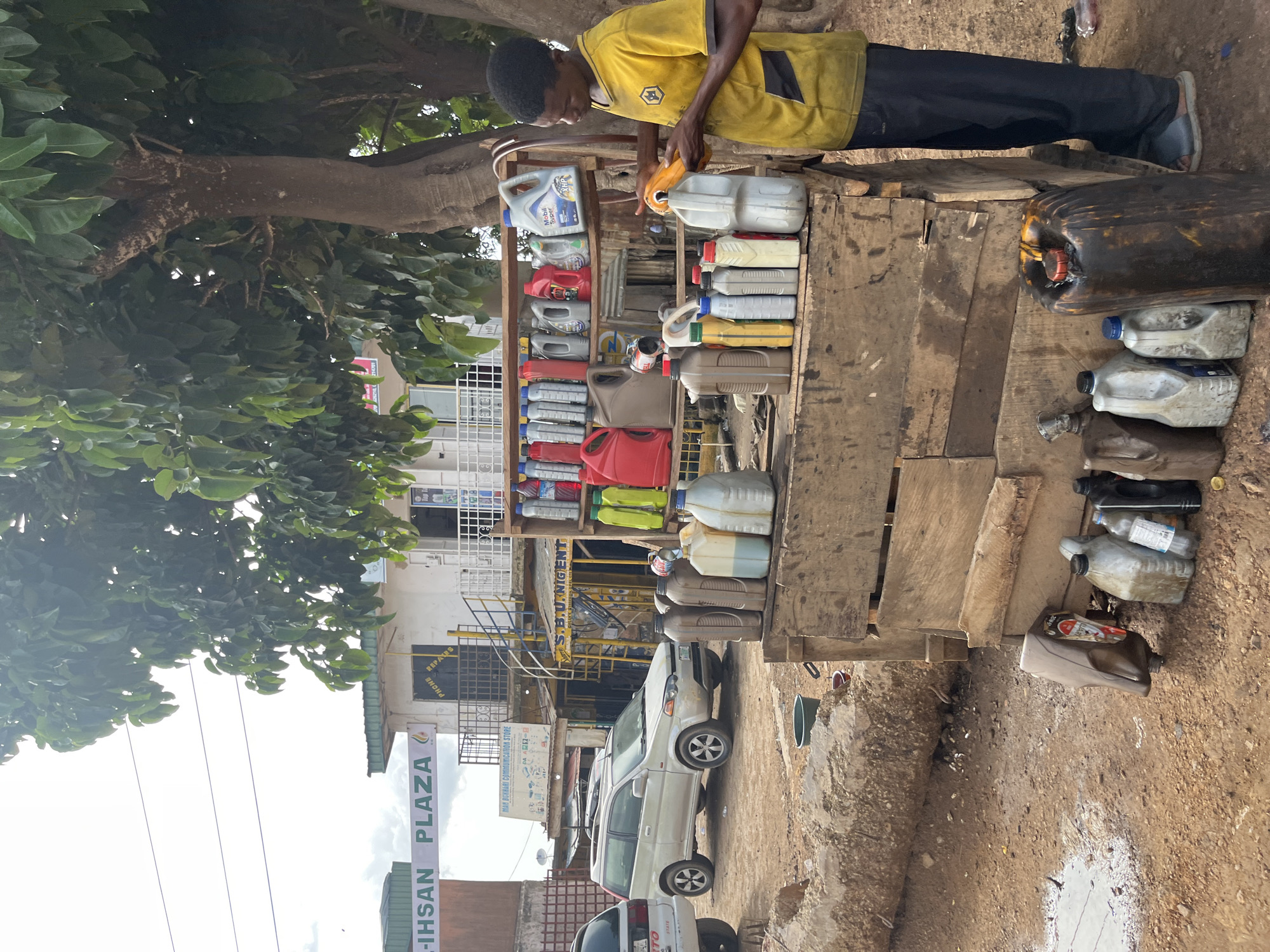 A man in a yellow shirt and black pants stands at a roadside stall, tending to a collection of plastic containers filled with liquids, likely motor oil or fuel. The stall is constructed from rough wooden planks, with containers stacked on shelves and on the ground. The scene is outdoors, with a large tree providing shade over part of the stall. In the background, a building with signage for "IHSAN PLAZA" and "MAN BUKHARI COMMUNICATION STORE" is visible, along with a white van and other vehicles. The ground is dirt and gravel, suggesting a somewhat informal commercial area. The man appears focused on his task, and the overall atmosphere is that of a busy marketplace or roadside service point. The lighting suggests it is daytime. Visible text includes brand names like "Mobil Super" and store names.