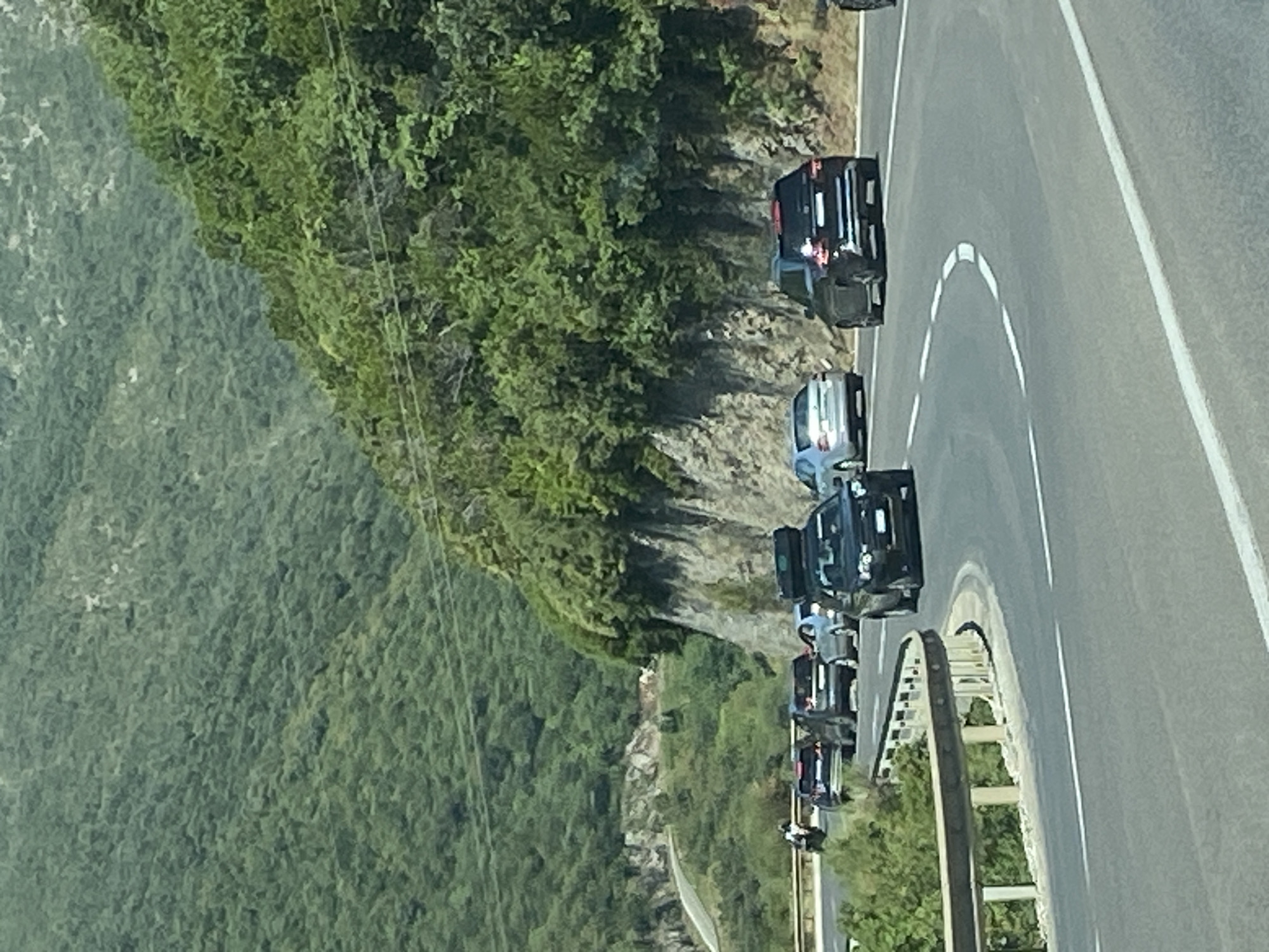 In Prijevor, Montenegro, a traffic jam occurs on a winding road. Several cars are visible on the asphalt road, with a black SUV in the foreground carrying luggage on its roof. The road is flanked by a steep, rocky mountainside covered in dense green foliage. The sky is bright, suggesting daytime.