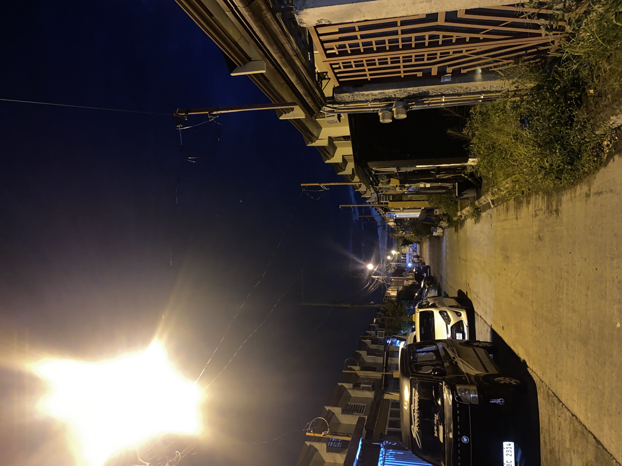The image depicts a street in a residential area of Davao City, Philippines at night. The sky is dark blue, indicating it is either early evening or late night. A bright street light illuminates the scene from the left, casting long shadows. On the left side of the street, a row of houses with similar architecture, featuring beige exteriors and angled rooflines, are visible. Several cars are parked along the street, including a black van in the foreground with license plate "BC 2314". Further down the street, more cars are parked, and street lights can be seen. On the right side of the street, another row of houses with beige exteriors is visible, along with a metal gate and a concrete wall. Overgrown grass and plants line the sidewalk on the right. Utility poles and electrical wires are present throughout the scene, common in urban Filipino landscapes. The overall impression is a quiet residential street at night, illuminated by artificial light.
