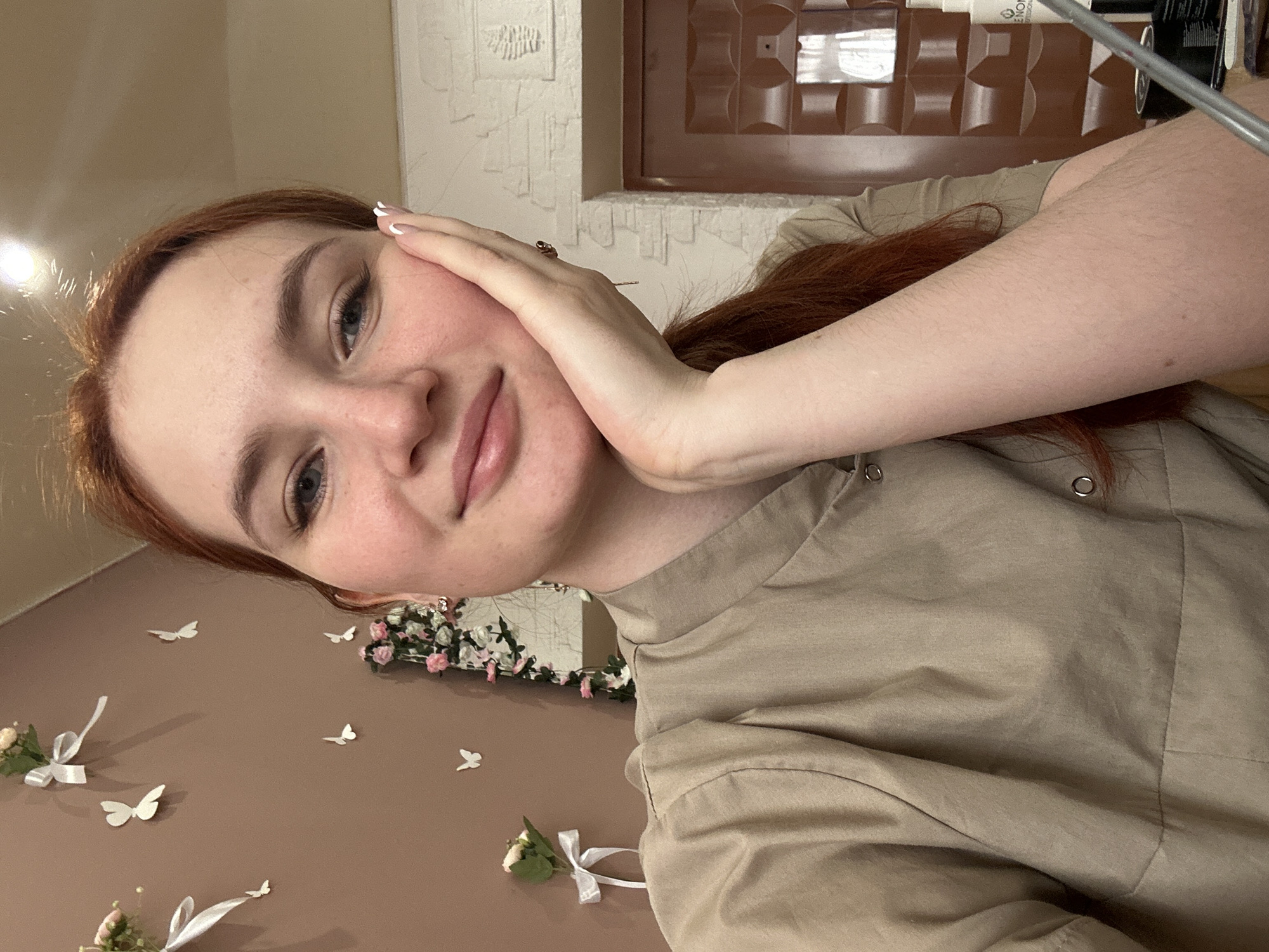 A close-up selfie of a fair-skinned person with reddish-brown hair and light-colored eyes. They are wearing a tan-colored collared shirt or uniform and resting their hand on their cheek. The background features a tan wall adorned with white paper butterflies and small floral arrangements with white ribbons. A mirror with a floral garland is partially visible. Above the wall is textured white paint or wall covering. A dark brown door or cabinet is located above that. The location is suggested to be Chaikovsky, Russia, but there is no definitive element in the image to confirm this.