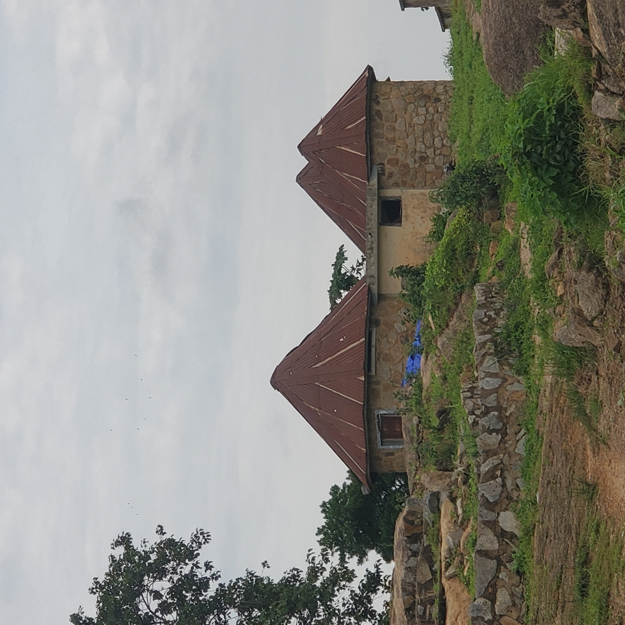 The image depicts a rural landscape in the city of Jos, Nigeria, featuring two circular, stone-built structures with conical, corrugated iron roofs. These structures are situated on a rocky incline amidst lush green vegetation. In the foreground, a stone wall built with large, irregular stones delineates a path or garden area. The sky is overcast with scattered clouds, and a few birds are visible flying in the distance. To the left of the structures, a large tree with dense foliage stands prominently, adding to the natural setting. The overall scene suggests a peaceful, traditional dwelling or perhaps a historical site within the Nigerian landscape.