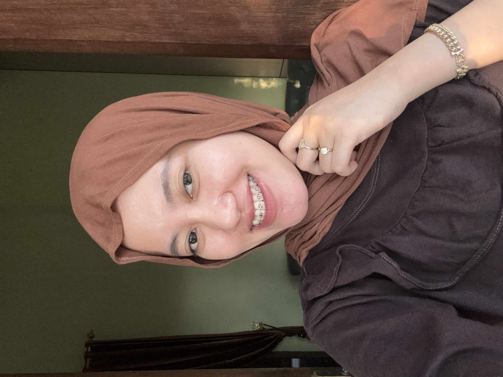 In this close-up, self-taken photograph, a young woman is captured from the chest up, looking directly at the camera. She is wearing a brown hijab that drapes around her head and neck, and a dark brown, ruffled shirt. Her teeth are adorned with silver braces, and she is smiling. On her left hand, she wears two rings: one on her index finger and another on her middle finger. A gold bracelet is visible on her right wrist. The background is dimly lit, with a dark brown door frame on the right and a dark curtain on the left. The wall behind her is a muted green. The lighting suggests it might be indoors with natural light coming from the right, possibly indicating a domestic setting in Gadingrejo, Indonesia.