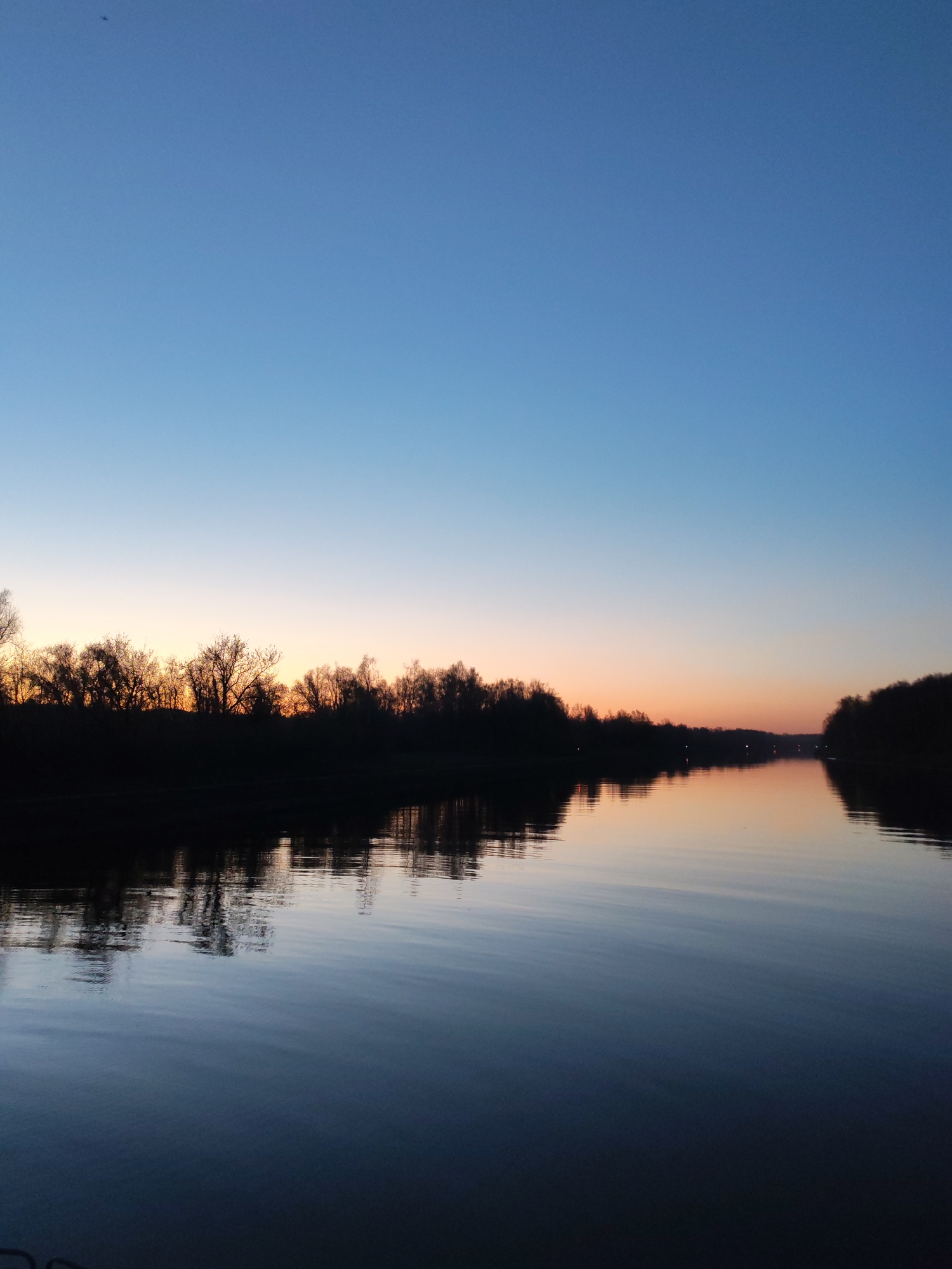 The image depicts a calm river at dusk in Yakhroma, Russia. The sky transitions from a deep blue at the top to a lighter blue-orange near the horizon. Silhouetted trees line both banks of the river. The water reflects the colors of the sky and the dark shapes of the trees, creating a mirror-like effect. There are no visible people or objects in the water or on the shore. The scene conveys a sense of tranquility.