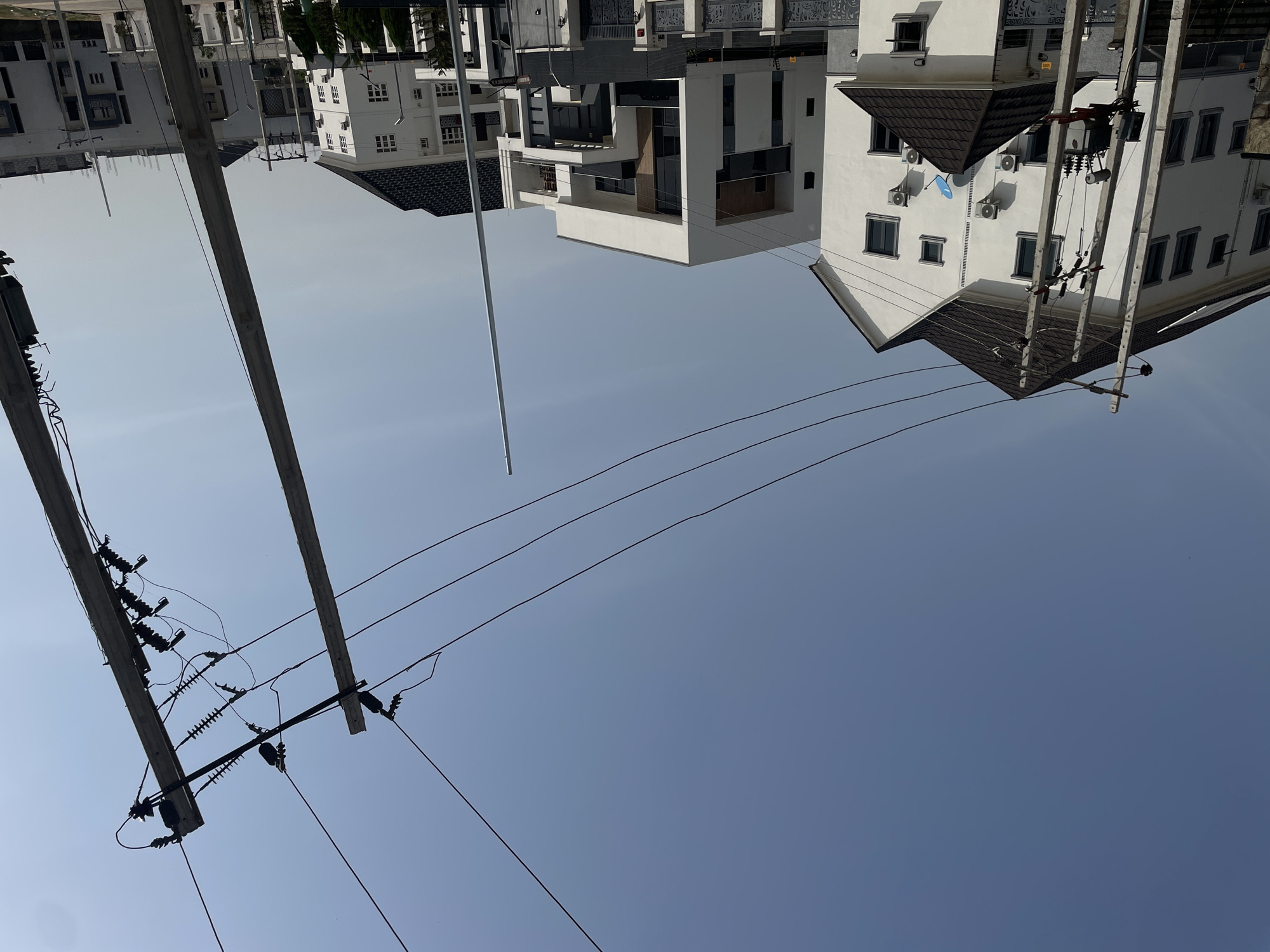 This image, captured in Gwarinpa, Nigeria, presents an inverted view of an urban or suburban street scene under a clear, bright sky. The sky dominates the lower and middle portions of the frame, transitioning from a light blue at the bottom to a slightly darker shade higher up, indicating daytime with no clouds.
In the foreground, prominent electrical utility poles with multiple cross-arms, numerous insulators, and a dense network of power lines and cables are visible. These poles appear to extend upwards from the "bottom" of the frame (which is actually the top if the image were correctly oriented), silhouetted against the sky. The wiring shows a complex system of distribution.
Above the electrical infrastructure, in what would be the background if oriented correctly, several modern, multi-story buildings are depicted upside down. These structures feature predominantly white or light-colored facades, often with dark-tiled, peaked roofs. Many buildings display multiple windows, some with dark frames, and some include balconies with railings. Air conditioning units are visible on the exterior walls of several buildings, and one building features a distinct blue, possibly parabolic, object (likely a satellite dish or a component of an AC unit). The architectural style suggests contemporary residential or commercial properties.
No people are visible, and no specific activity is taking place; the scene is static. The overall impression is of a sunny day. No discernible text is visible on any of the structures or utility components. The unique, inverted perspective is the most striking visual aspect of the media file.