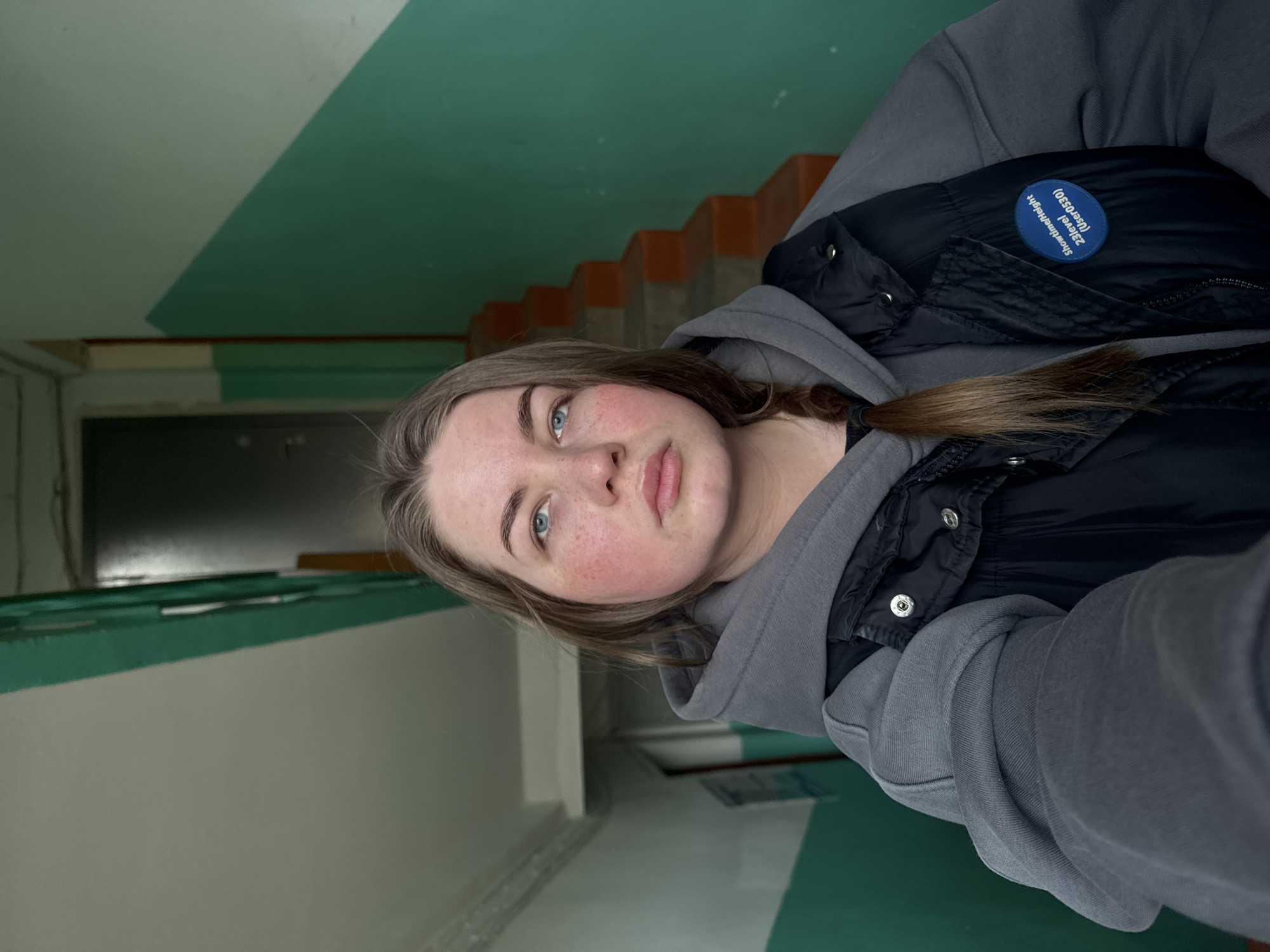The image is a rotated selfie of a young woman with fair skin, blue eyes, and light brown hair. Her cheeks are visibly flushed with rosy red color. She is wearing a gray hoodie and a black puffer vest. The vest has a blue round patch with white writing on it, visible on the upper right side of the frame. Her hair is parted and braided on one side.
The background appears to be an interior hallway or stairwell. The walls are painted with a light green stripe at the top and a white stripe below. There are orange stairs visible in the upper part of the image. A black door is seen in the distance to the left.
Based on the location (Chaikovsky, Russia), the image likely depicts an interior space in a multi-story building such as an apartment complex or public building.