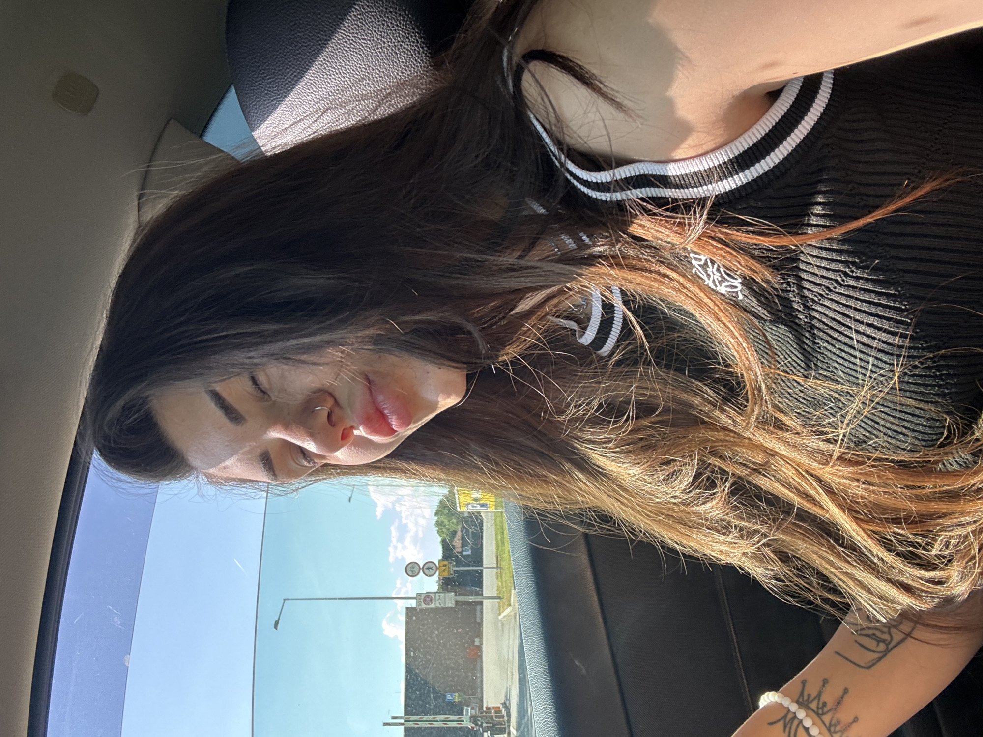 A young woman with long, brown hair, wearing a black ribbed top with white trim, is seen inside a car. She is looking to her right with her eyes closed and lips slightly parted, with sunlight illuminating her face. Her left arm, adorned with a bracelet and tattoos, is visible. Outside the car window, a street scene is visible with a traffic light, signs, a building, and trees under a clear blue sky. The interior of the car shows a dark-colored seat and part of the ceiling. The lighting suggests it is daytime. There are no visible indications of the specific location within Sofia, Bulgaria.