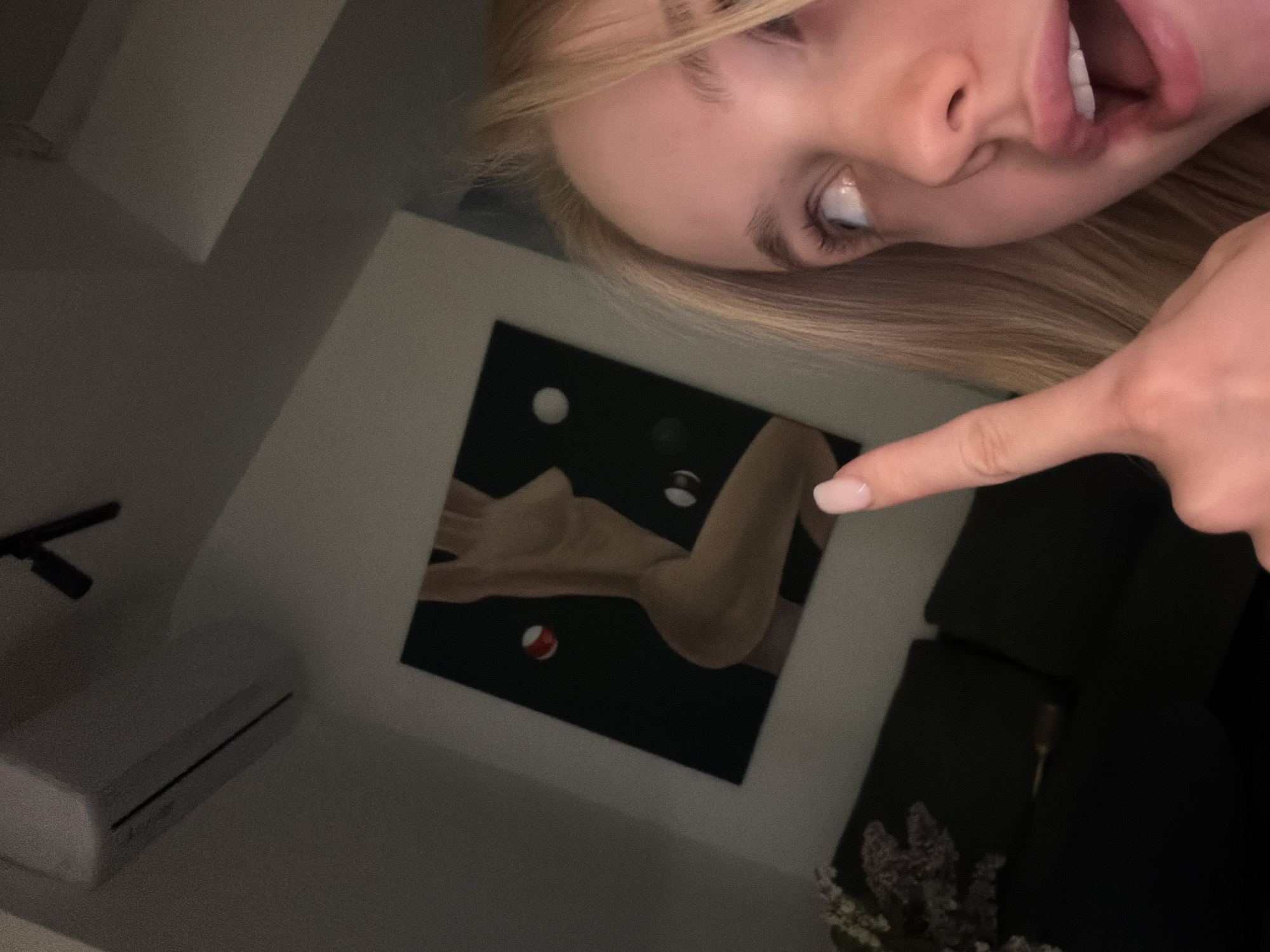 A blond-haired person points to a black-framed piece of art depicting a pair of arms and some balls, mounted on a white wall in a room with an air conditioning unit, light-colored ceilings and dark furniture. The location is Unknown, Unknown.