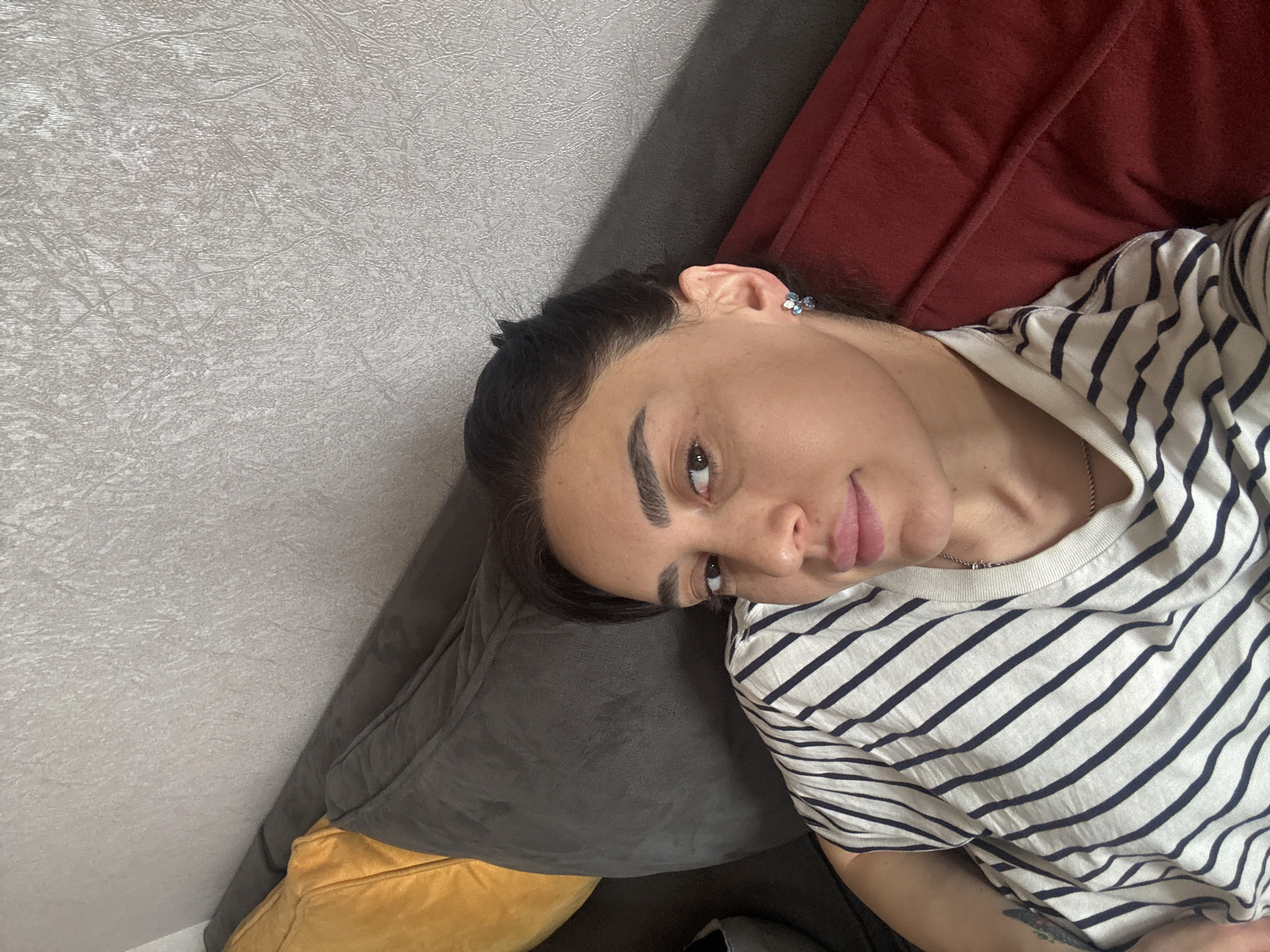 A woman is lying on a couch. The couch has grey, red, and yellow cushions. The wall behind her is textured and white. She is wearing a white and navy striped shirt and a butterfly earring. Her hair is pulled back. The location is not discernible from the image.