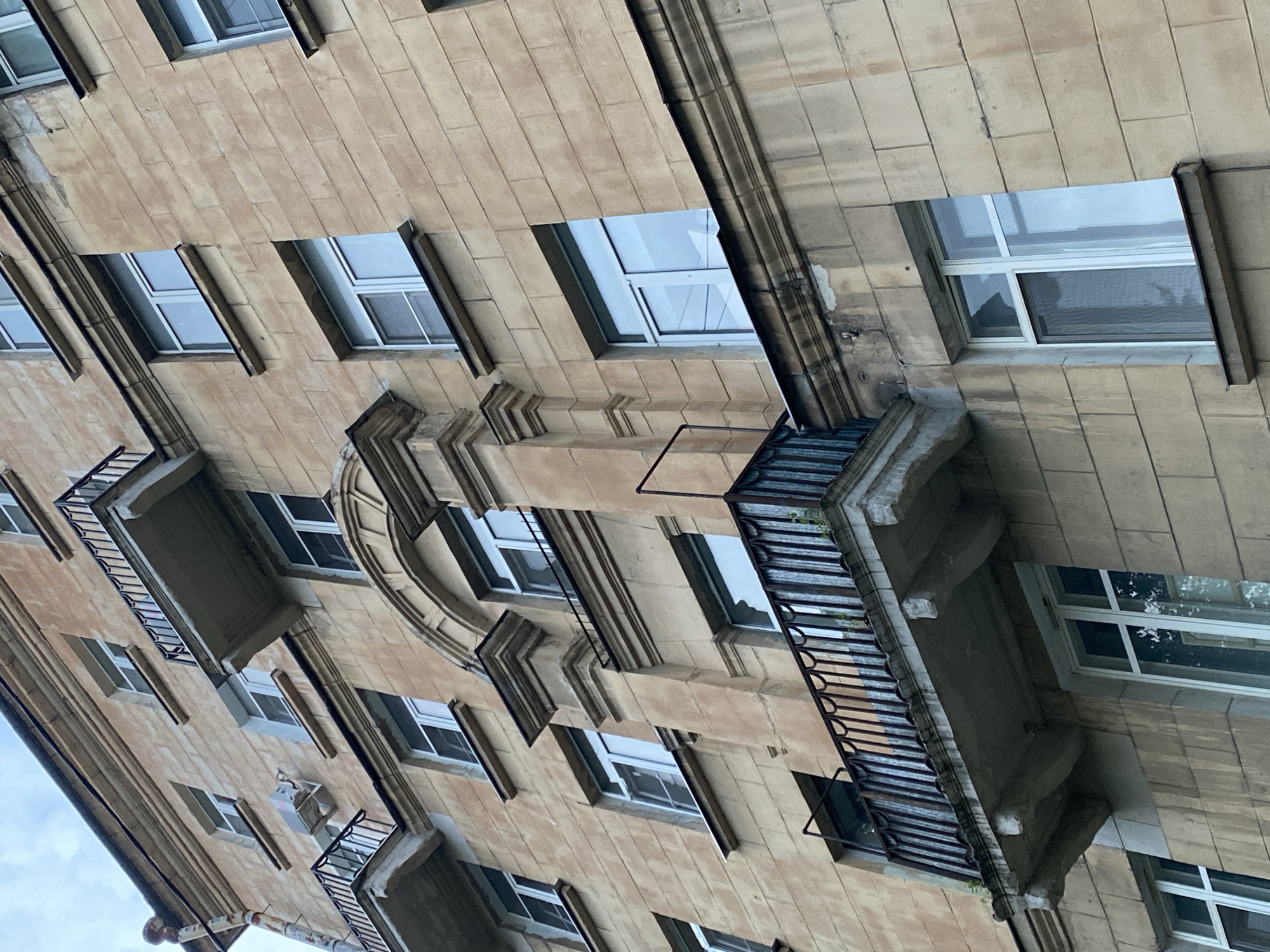 The image shows a close-up, low-angle view of the facade of an old, multi-story building in Saint Petersburg, Russia. The building is constructed of light brown, stone-like blocks, and features multiple windows, some with balconies. The architectural style suggests a pre-WWII construction, typical of many St. Petersburg residential buildings. The windows are mostly white-framed and appear to be double-glazed. Some balconies have ornate black metal railings. There is visible wear and tear on the facade, with some discoloration and chipped paint. An air conditioning unit is mounted on the wall near one of the upper windows. The sky is visible in the upper portion of the image and appears overcast. No people are visible in the image.