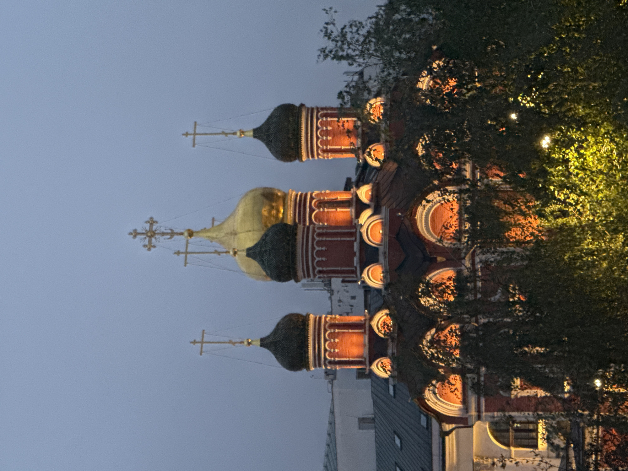 This is a medium-shot, eye-level, vertical photograph of a church in an urban environment at dusk. The church features multiple domes, one of which is golden and prominent in the center, flanked by smaller, darker domes. The building is adorned with red and white architectural details, and illuminated from within, casting an orange glow through arched windows. Trees with dense foliage are in the foreground, partially obscuring the lower part of the church. The sky is a muted blue-gray, characteristic of twilight. There are no people visible in the image. The overall scene conveys a sense of religious architecture in a city setting during the evening.